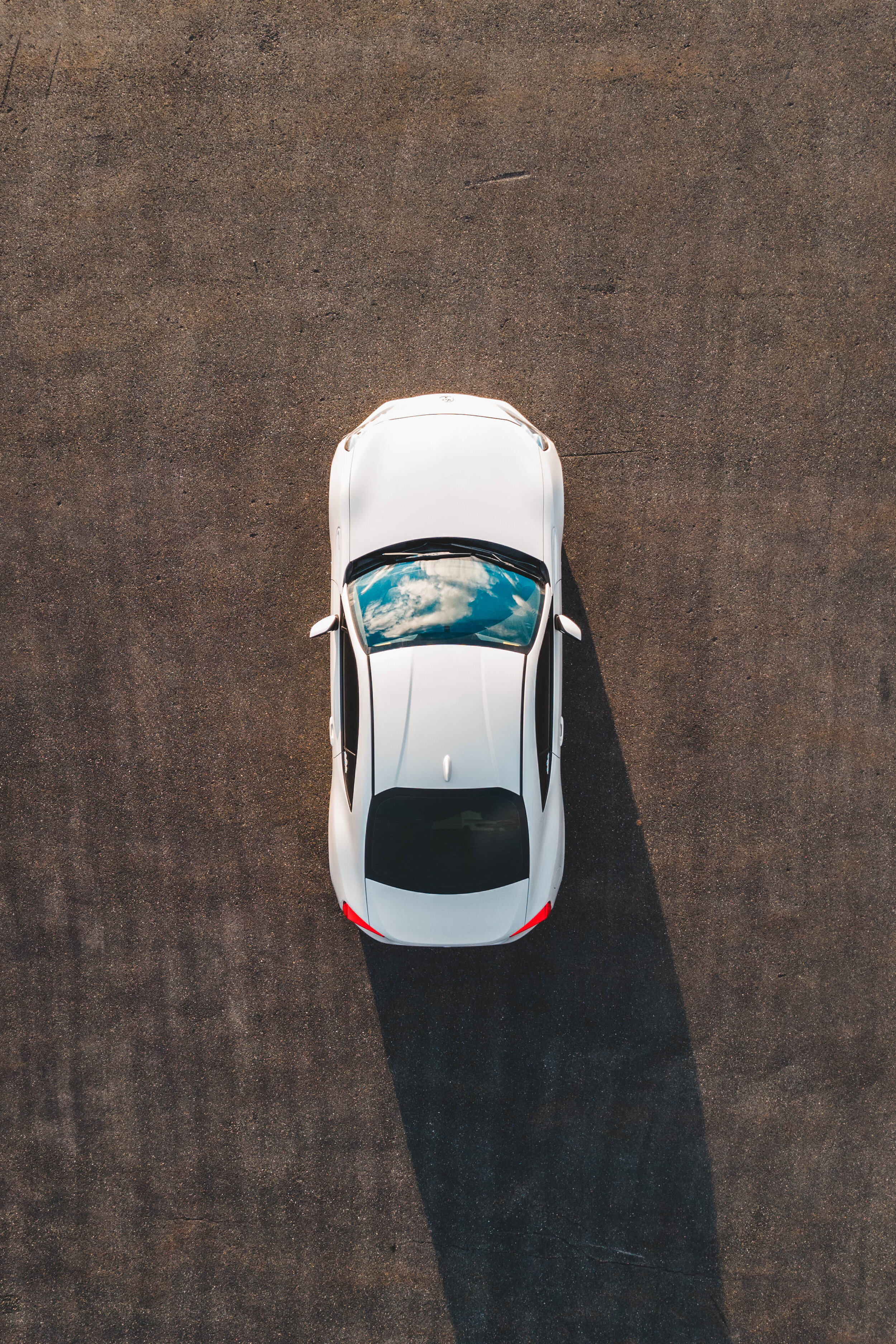 Aerial view of a white car parked on asphalt, reflecting clouds in its sunroof.