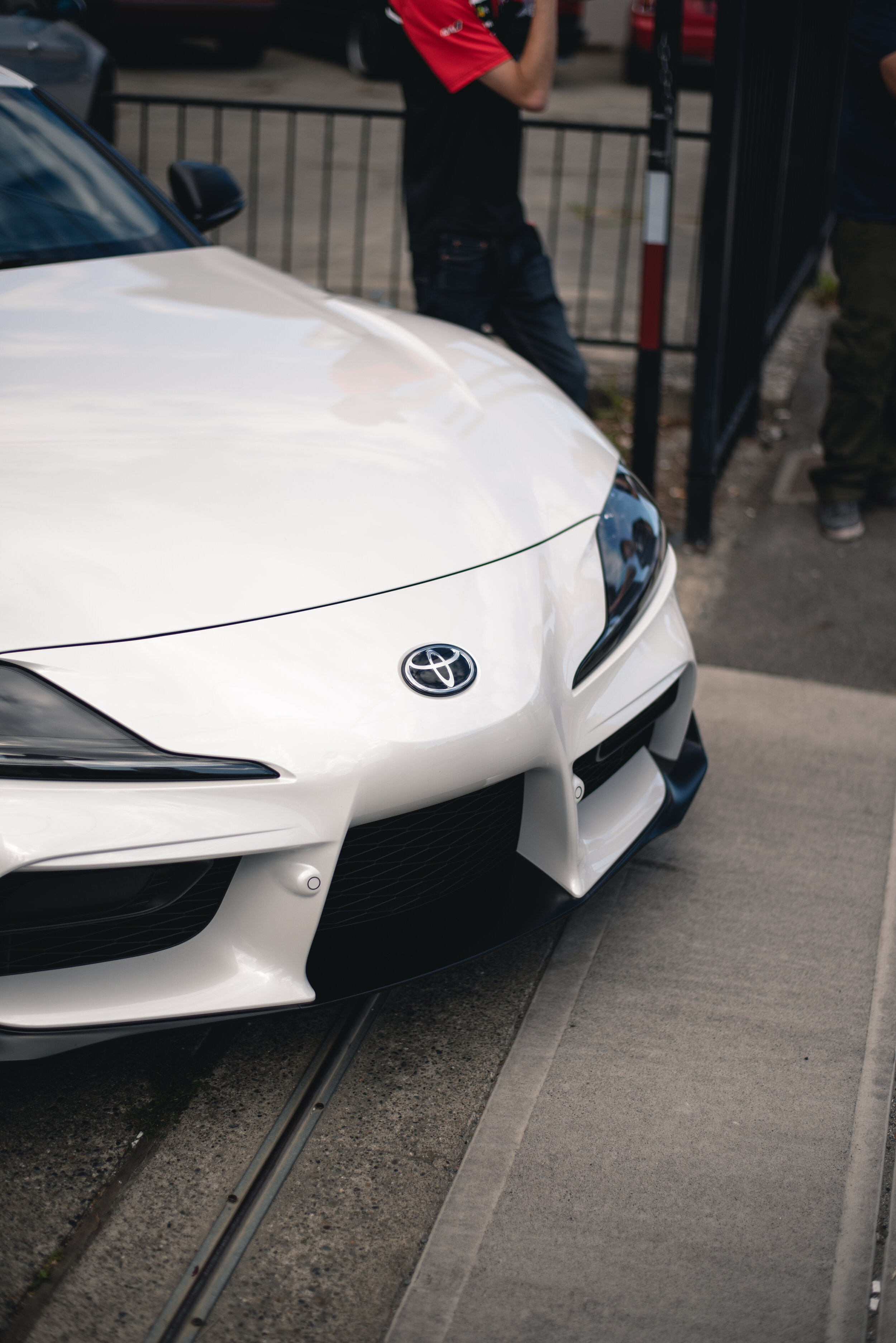 Front view of a white Toyota car with a visible logo and design details parked on a street with people nearby.