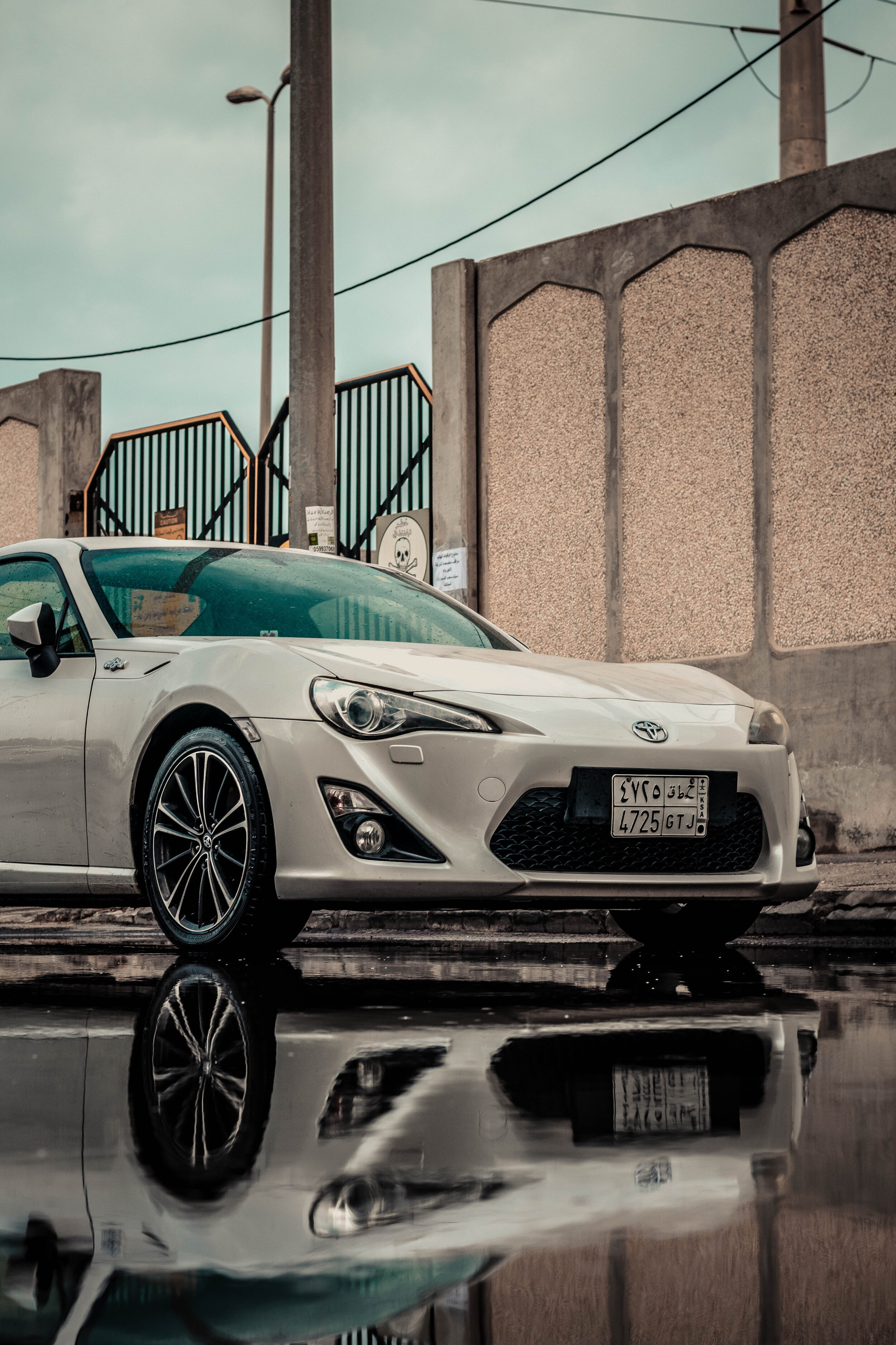 White sports car parked next to a reflective puddle, showcasing its reflection, with a background of a concrete wall and a gated entrance.