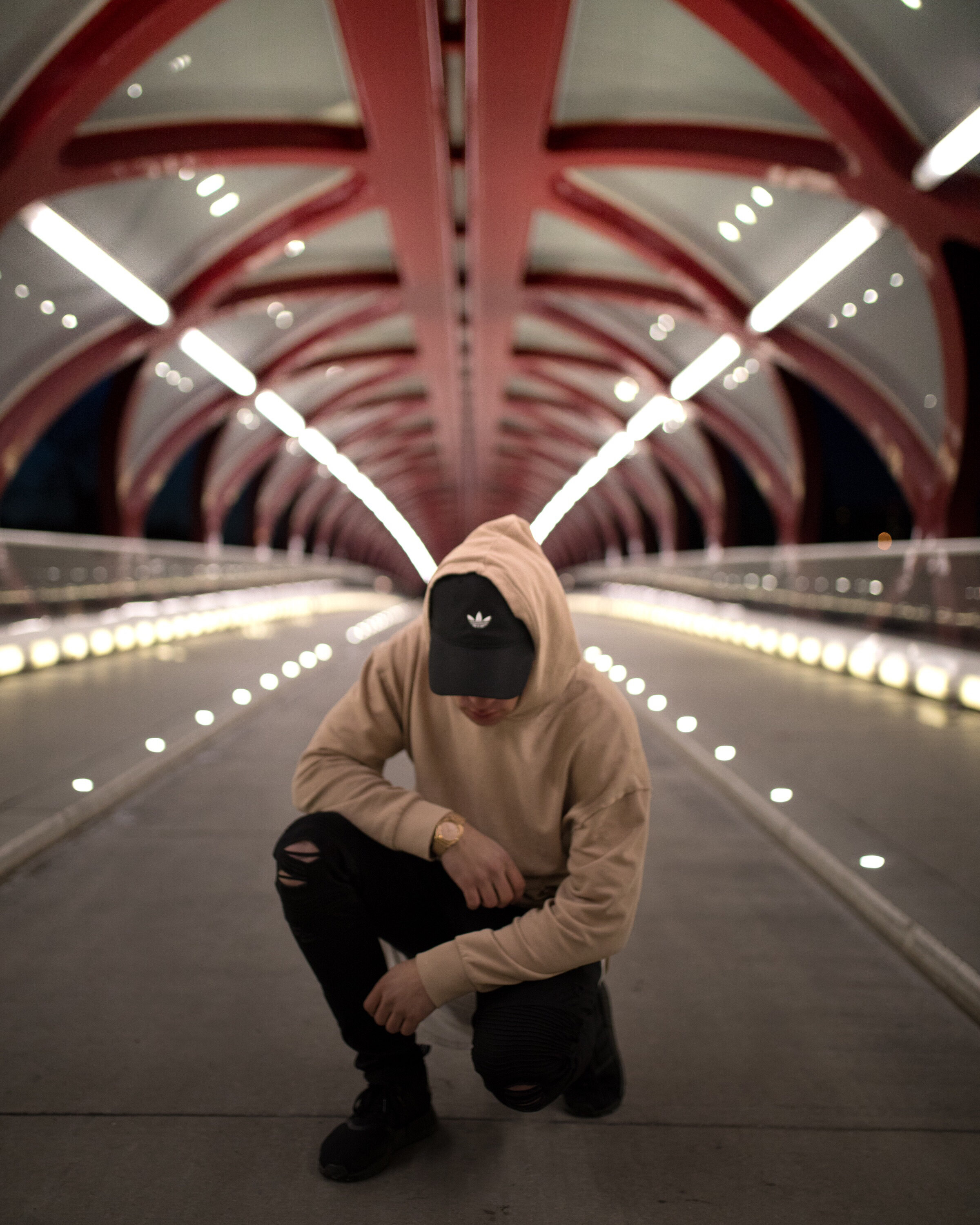 Person in hooded sweatshirt and cap crouching on illuminated bridge with arched metal structure.