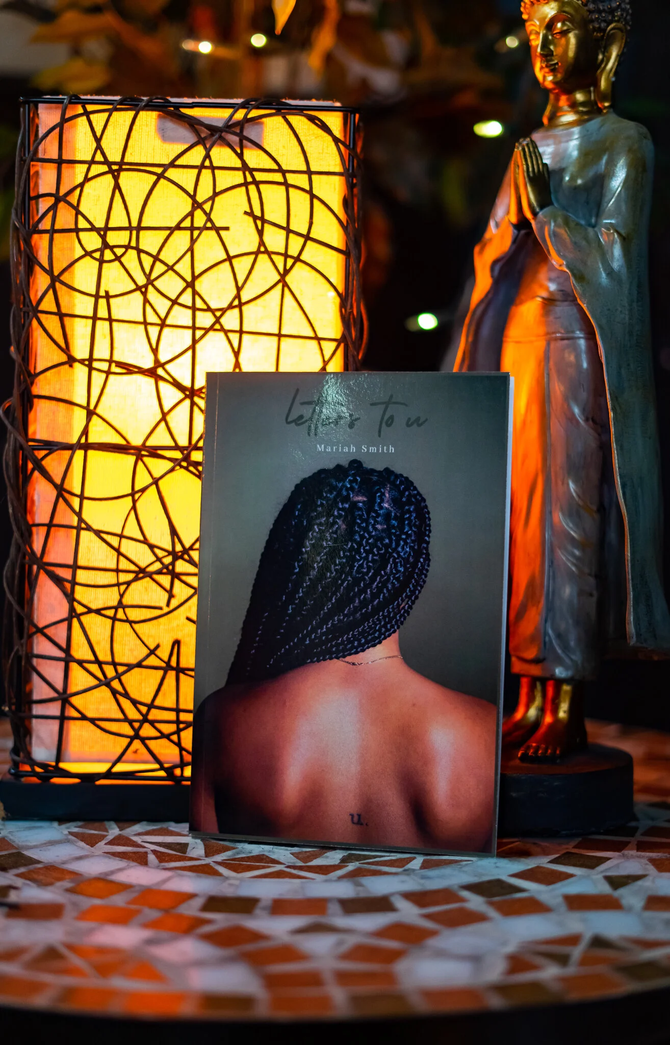 A book titled "Letter To U" by Mariah Smith features a back view of a woman with braided hair. The book is placed on a mosaic table next to a warm orange glowing lamp and a golden statue of Buddha with hands in prayer pose.