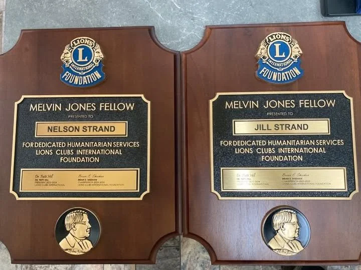 Two wooden plaques with Lions Club International Foundation logos, each awarded to Melvin Jones Fellow recipients Nelson Strand and Jill Strand for humanitarian services, with a profile of a person's face below each plaque.