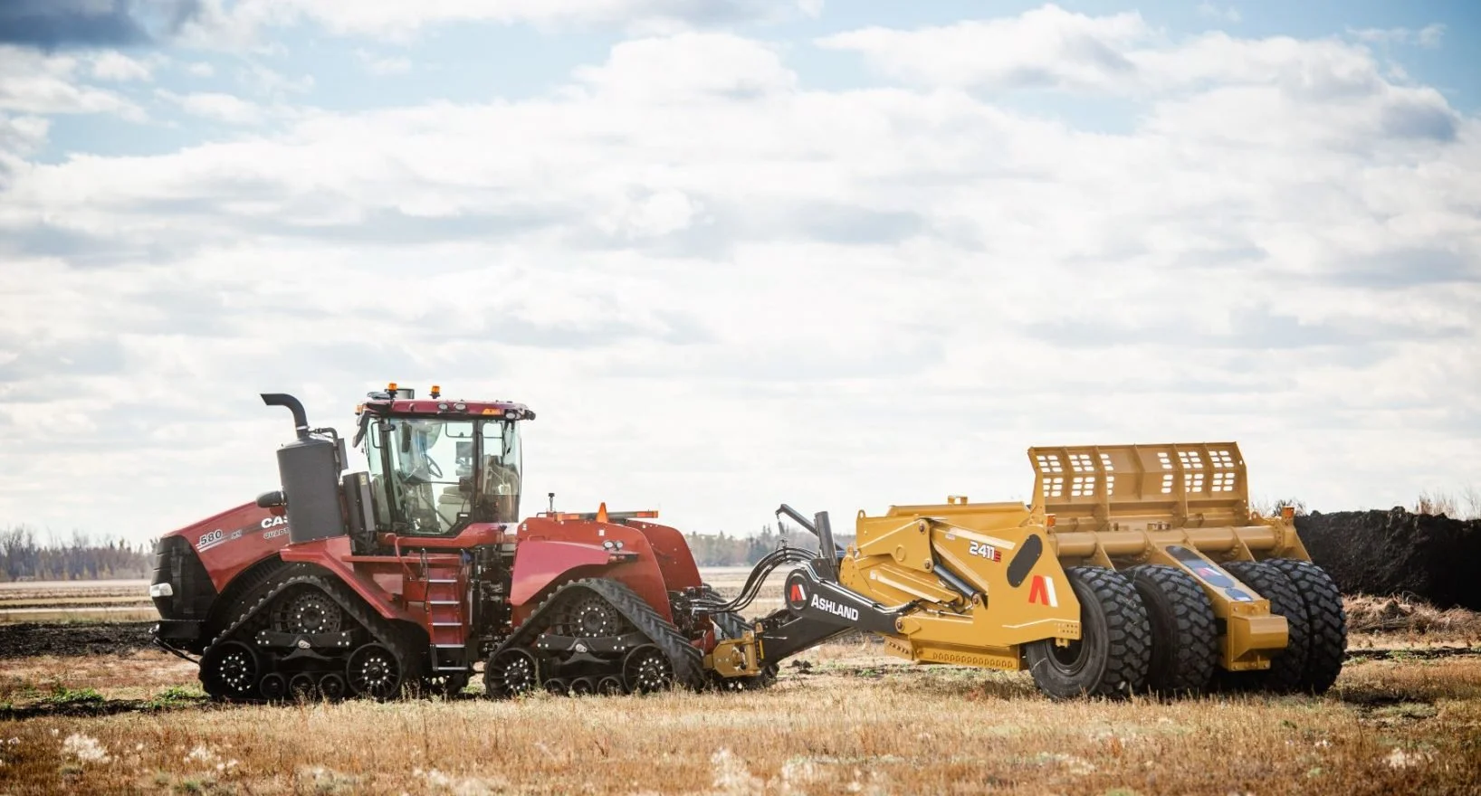 A red track tractor pulling a yellow soil compaction roller in a farmland field under a partly cloudy sky.