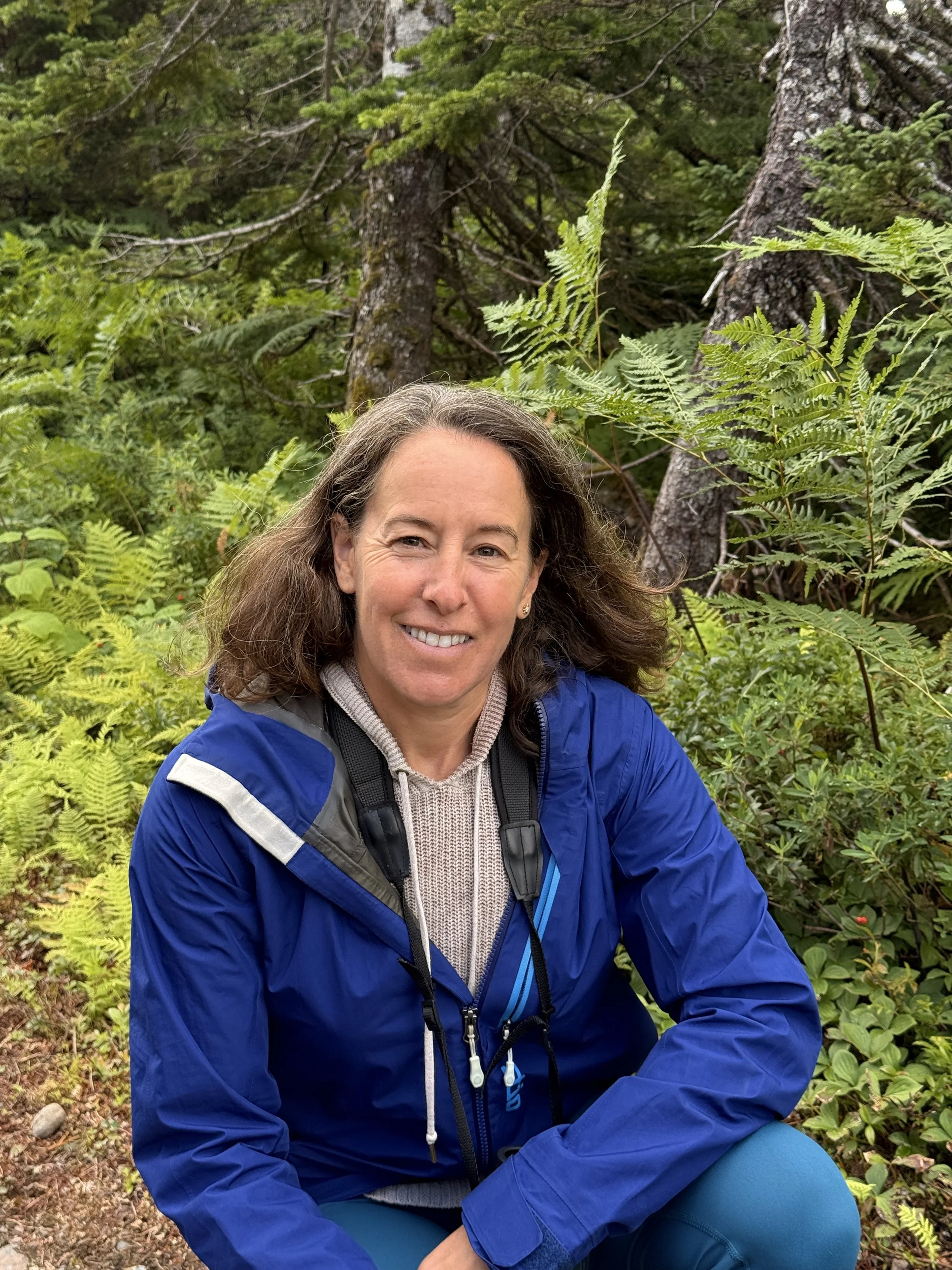 A woman with shoulder-length brown hair sitting outdoors in front of a rocky background, smiling at the camera