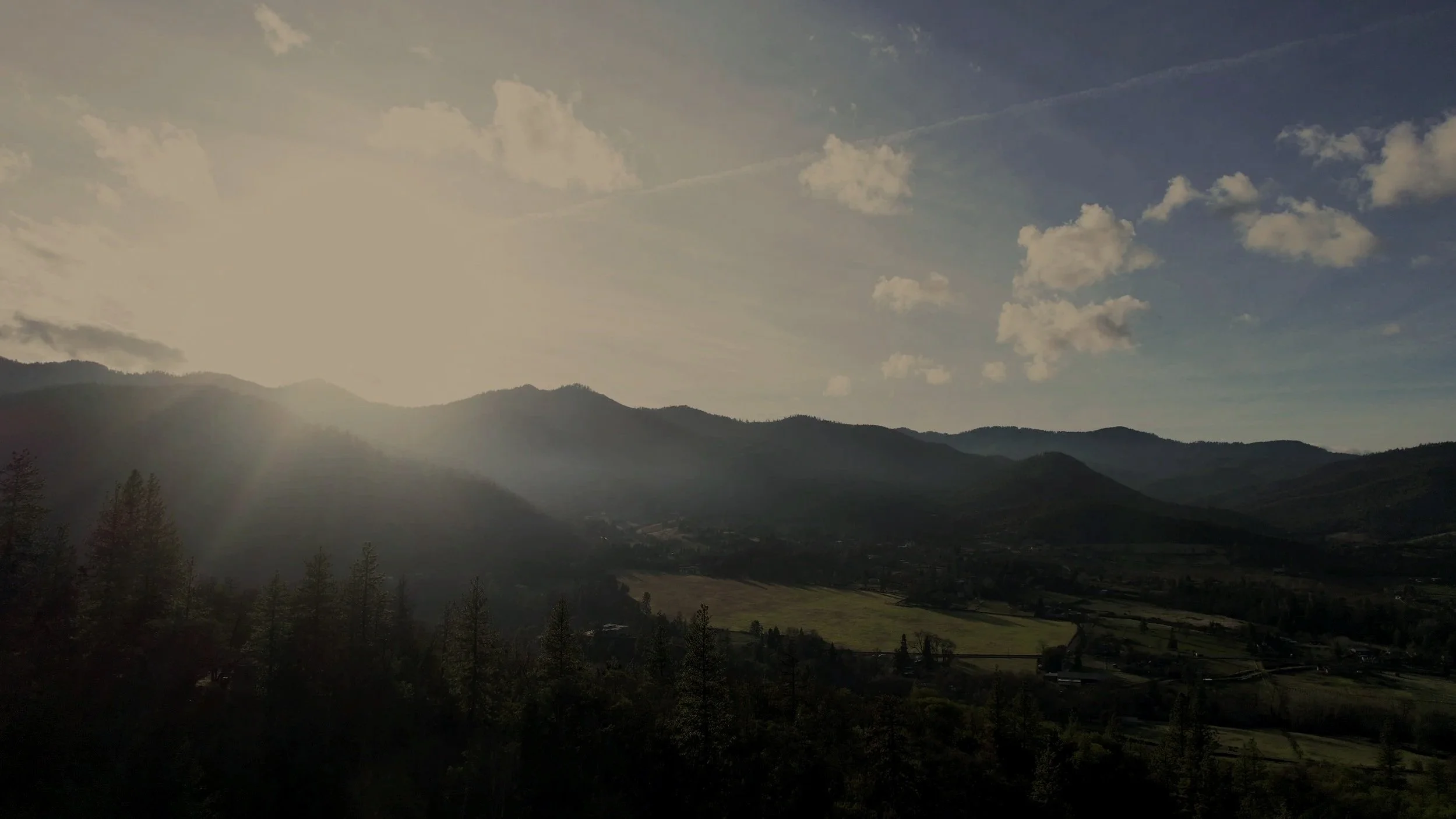Scenic view of a mountainous landscape with the sun shining through clouds, illuminating the rolling hills and green fields below.
