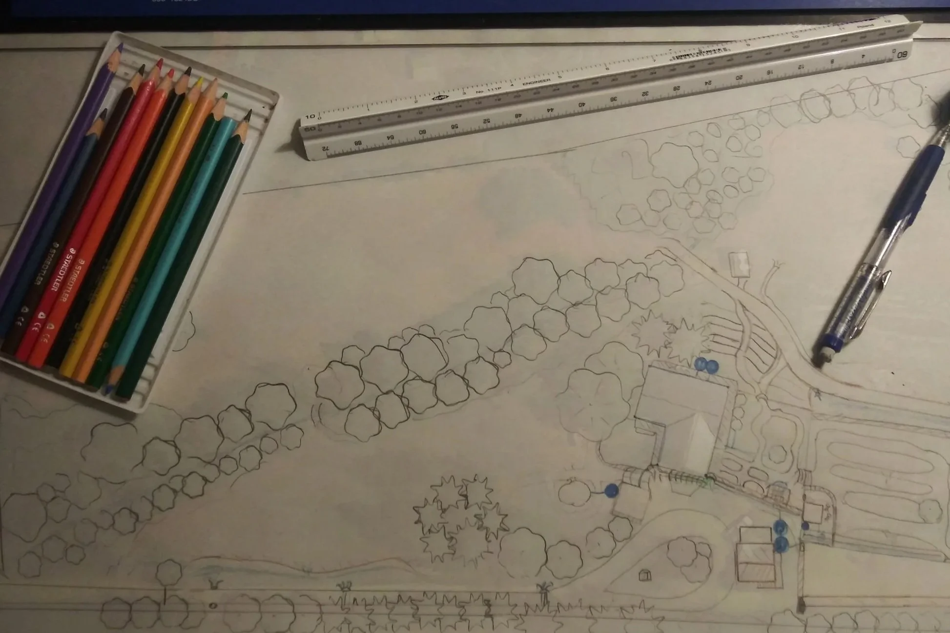 An architectural drawing of a landscape plan on paper with colored pencils, a ruler, and a pen placed around it.