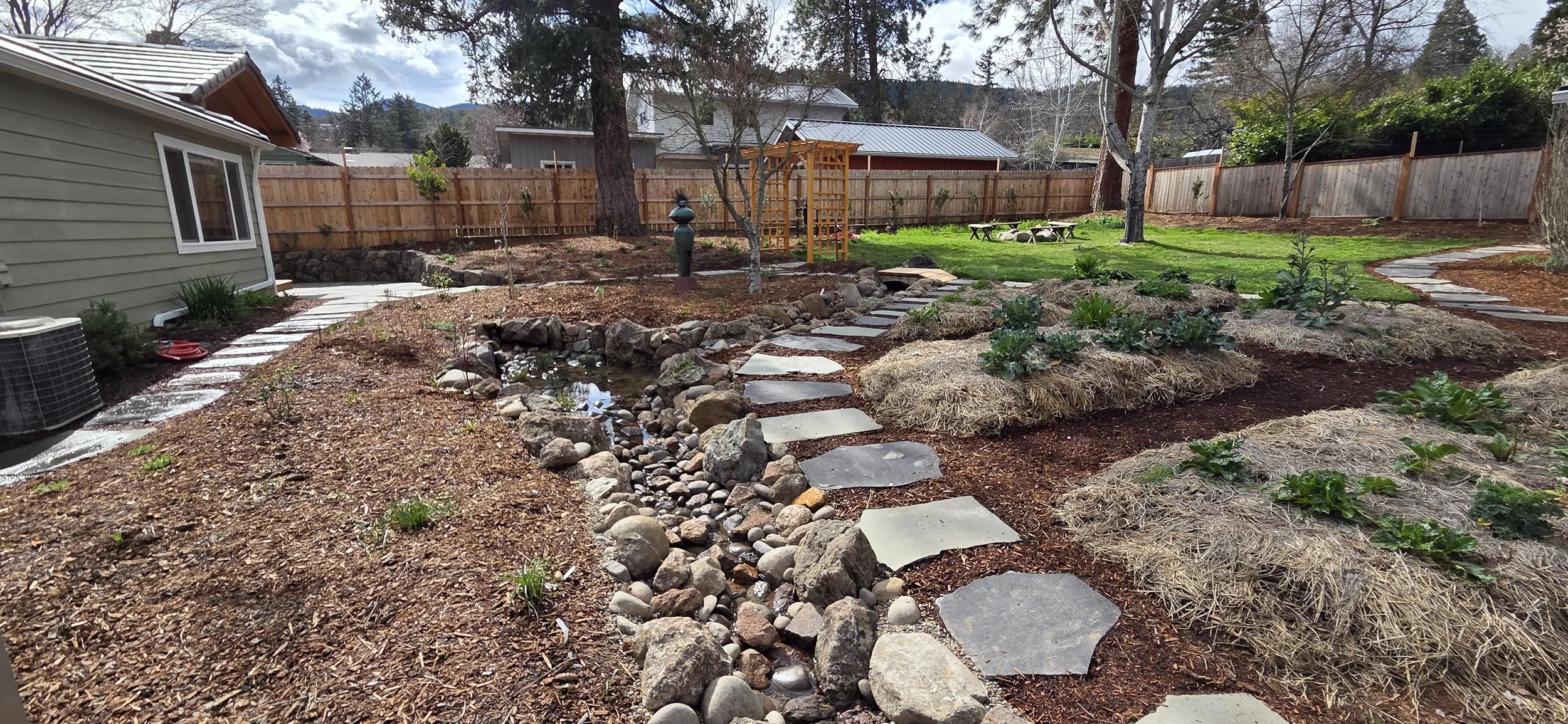 A backyard garden with a flagstone pathway, raingarden, small creek with rocks, veggie garden beds with green plants and straw mulch, green lawn, and wooden fence in the background.
