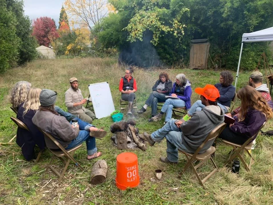 A group of people sitting in a circle outdoors, participating in a discussion or workshop near a campfire in a grassy area with trees and fall foliage in the background.