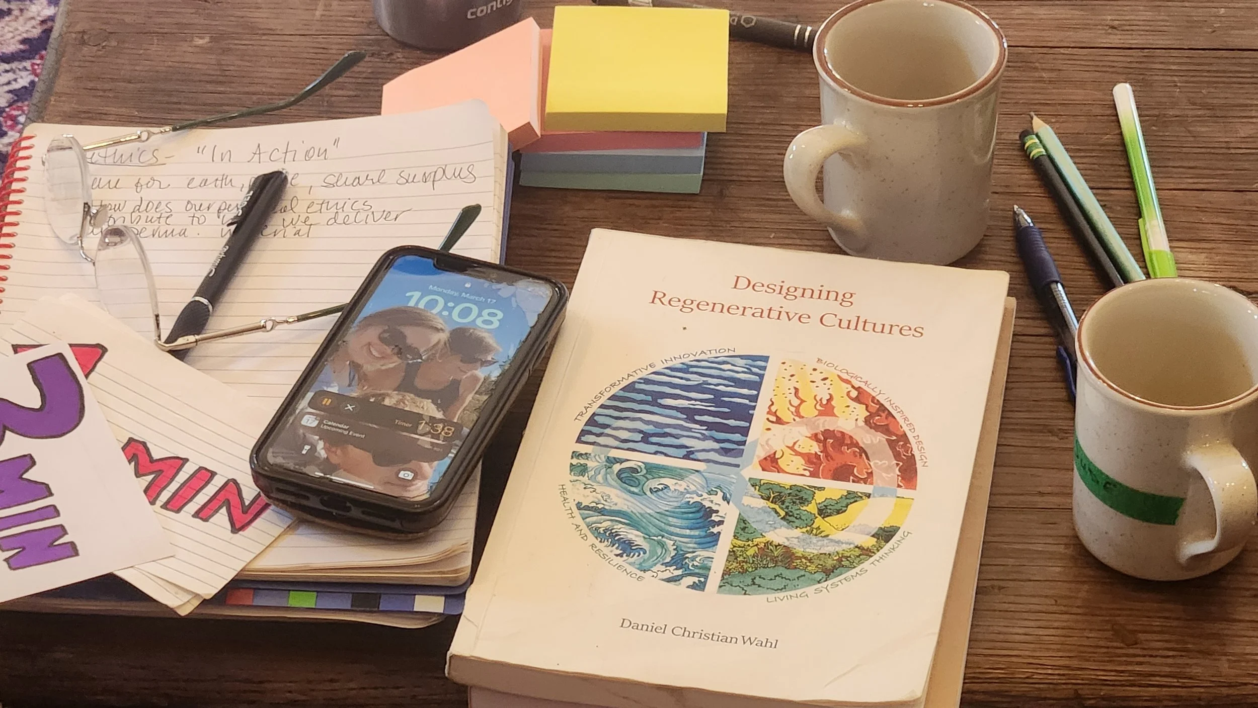 A desk with notebooks, pens, sticky notes, a photo of a young woman in sunglasses on a phone, a computer mouse, a book titled 'Designing Regenerative Cultures' by Daniel Christian Wahl, two mugs, and a colorful sign partially visible.