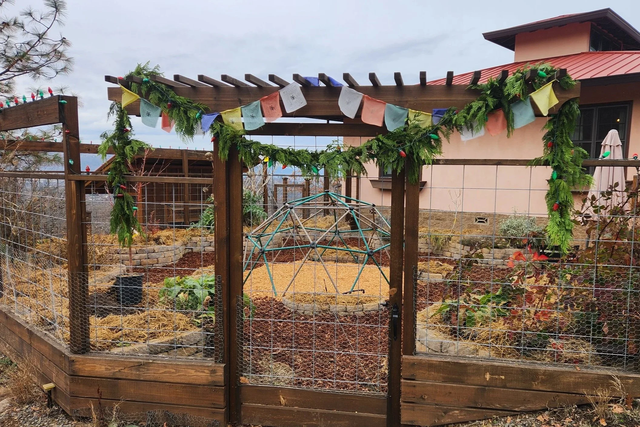 A permaculture kitchen garden enclosed by a wooden and wire fence, decorated with holiday garlands and colorful prayer flags, with a geometric garden structure and plants inside.