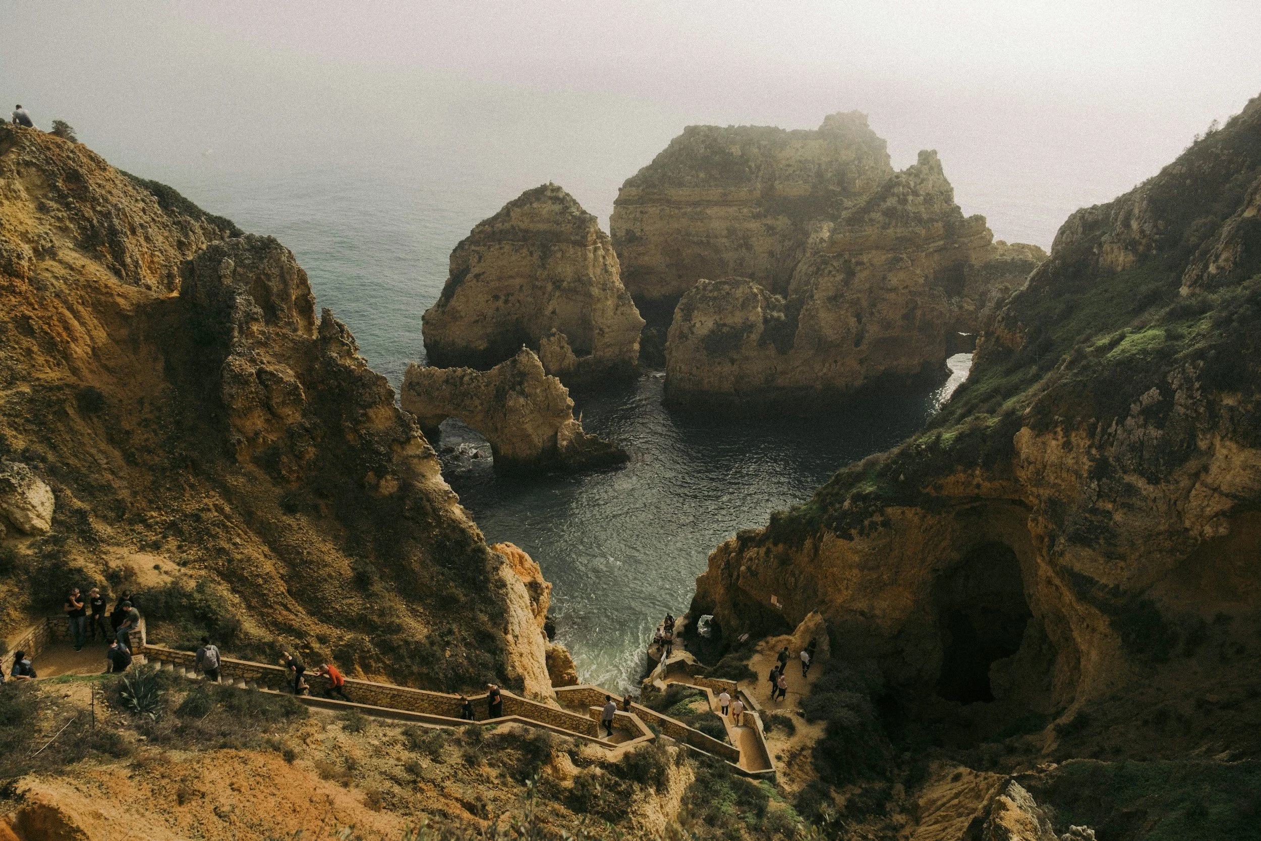 Coastal cliffs with large rock formations and a staircase leading down to the water, with visitors exploring the area.
