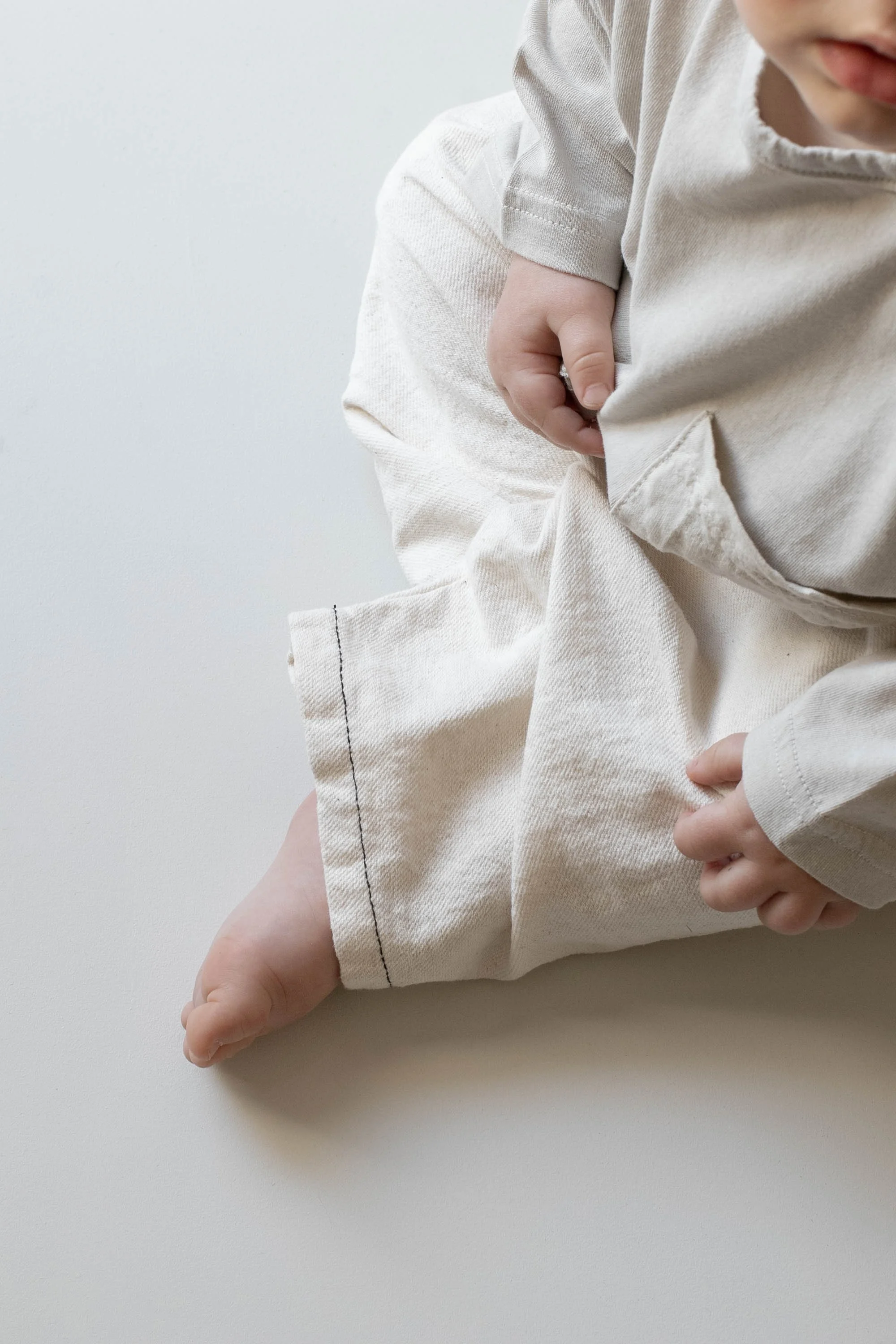 Close-up of a baby dressed in a beige outfit, sitting on a white surface with one leg stretched out and the other partially bent, with visible hands holding the pants.