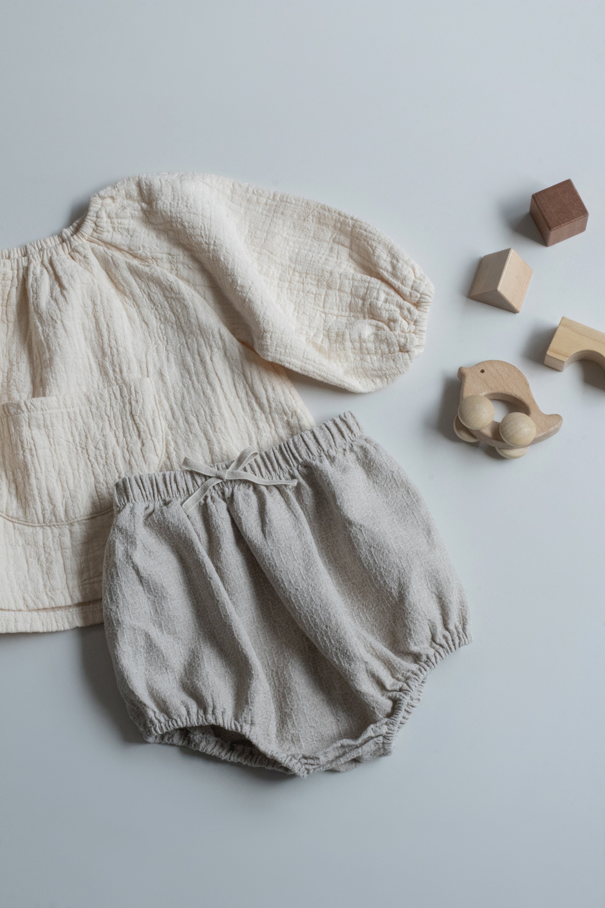 Clothing and toys for a baby, including a beige quilted jacket, gray bloomers, and wooden toy blocks and car on a white surface.