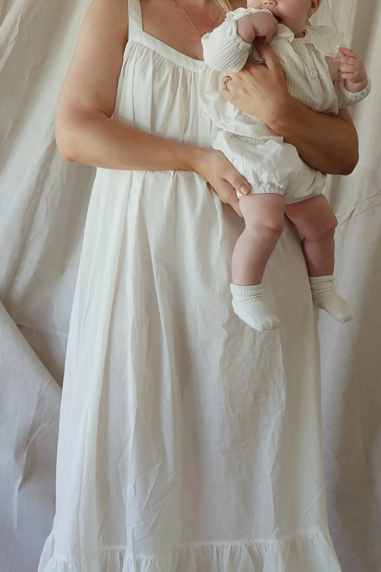 A woman dressed in a flowing, cream-colored dress holding a baby wearing a light-colored outfit with socks, against a neutral background.