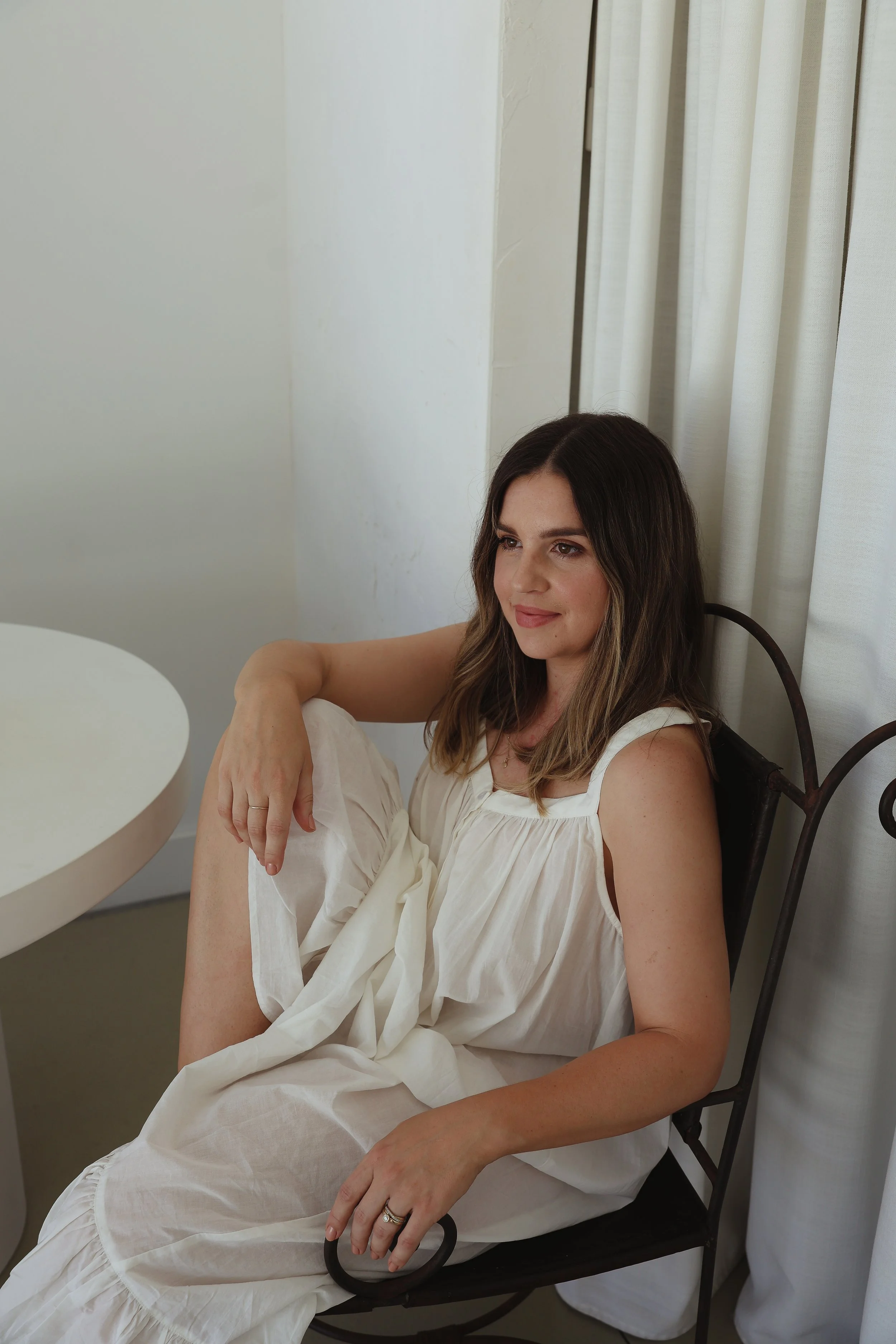A woman with long brown hair sitting on a black metal chair near white curtains, wearing a white sleeveless dress, with her right arm resting on her knee, smiling softly.