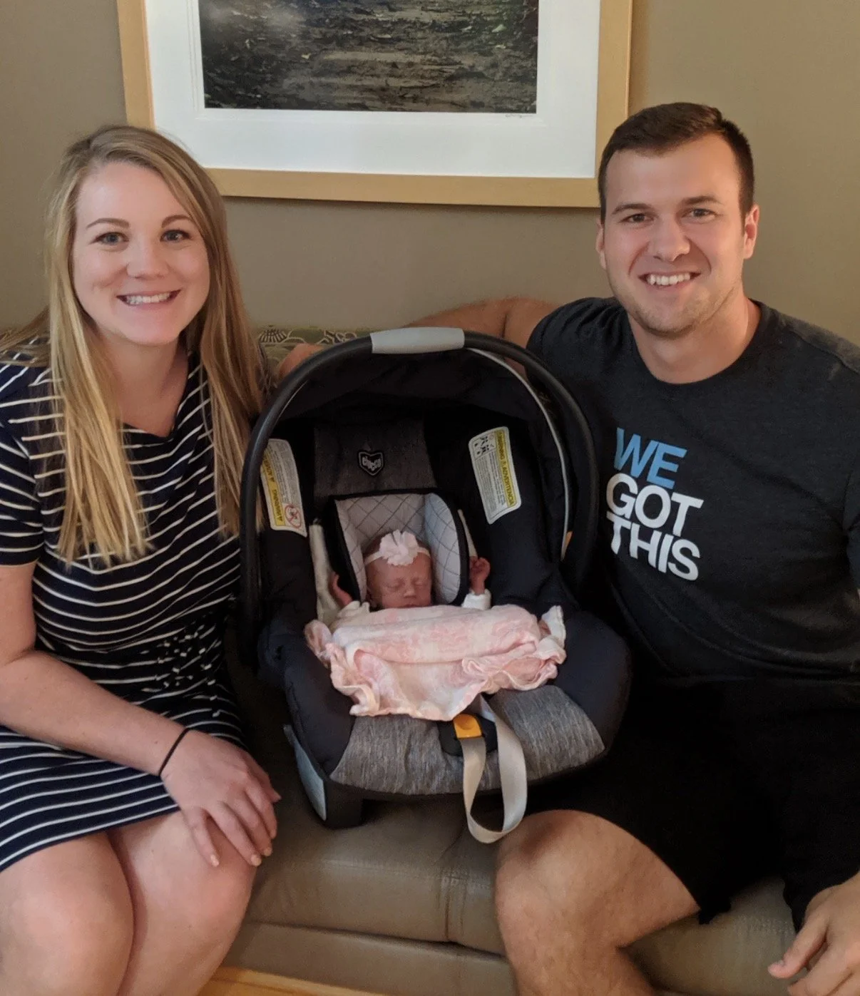 A happy couple sitting on a beige couch with their newborn baby in a car seat between them, in a room with a framed picture on the wall behind.