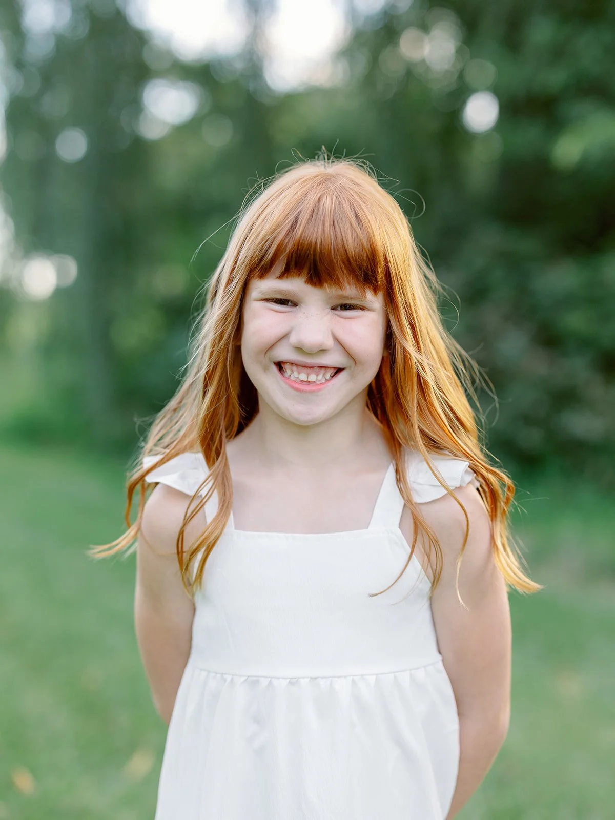 A young girl with red hair, smiling, wearing a white dress, outdoors with blurred greenery in the background.