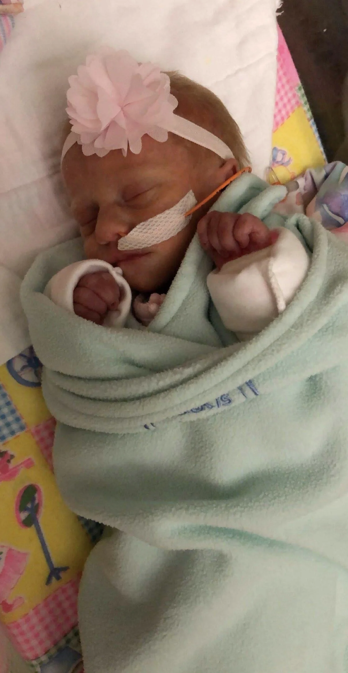 Newborn baby girl sleeping with a pink headband and flower, wrapped in a blanket, on a colorful quilt.