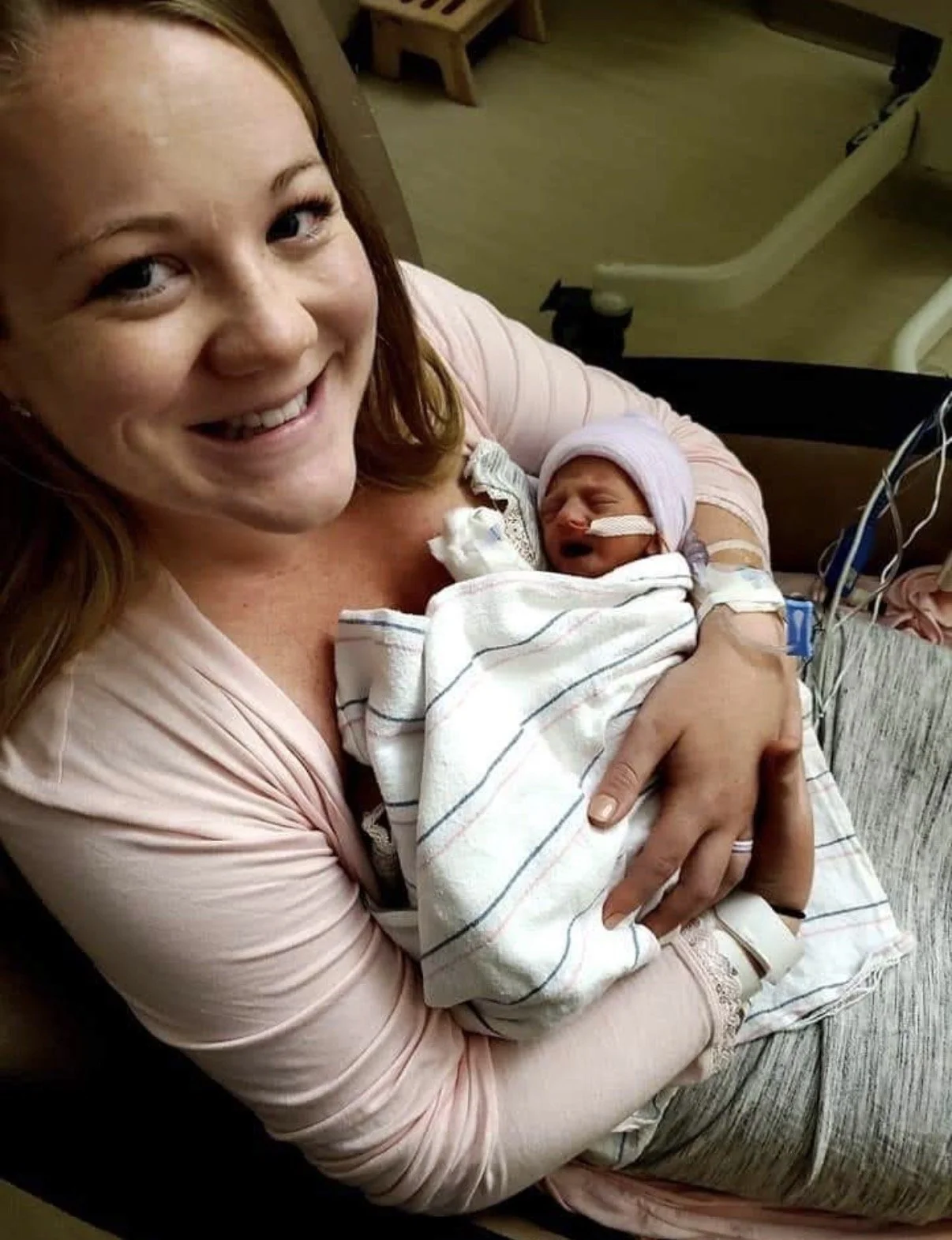 A woman with red hair holding a newborn baby wrapped in a hospital blanket, smiling in a hospital room.