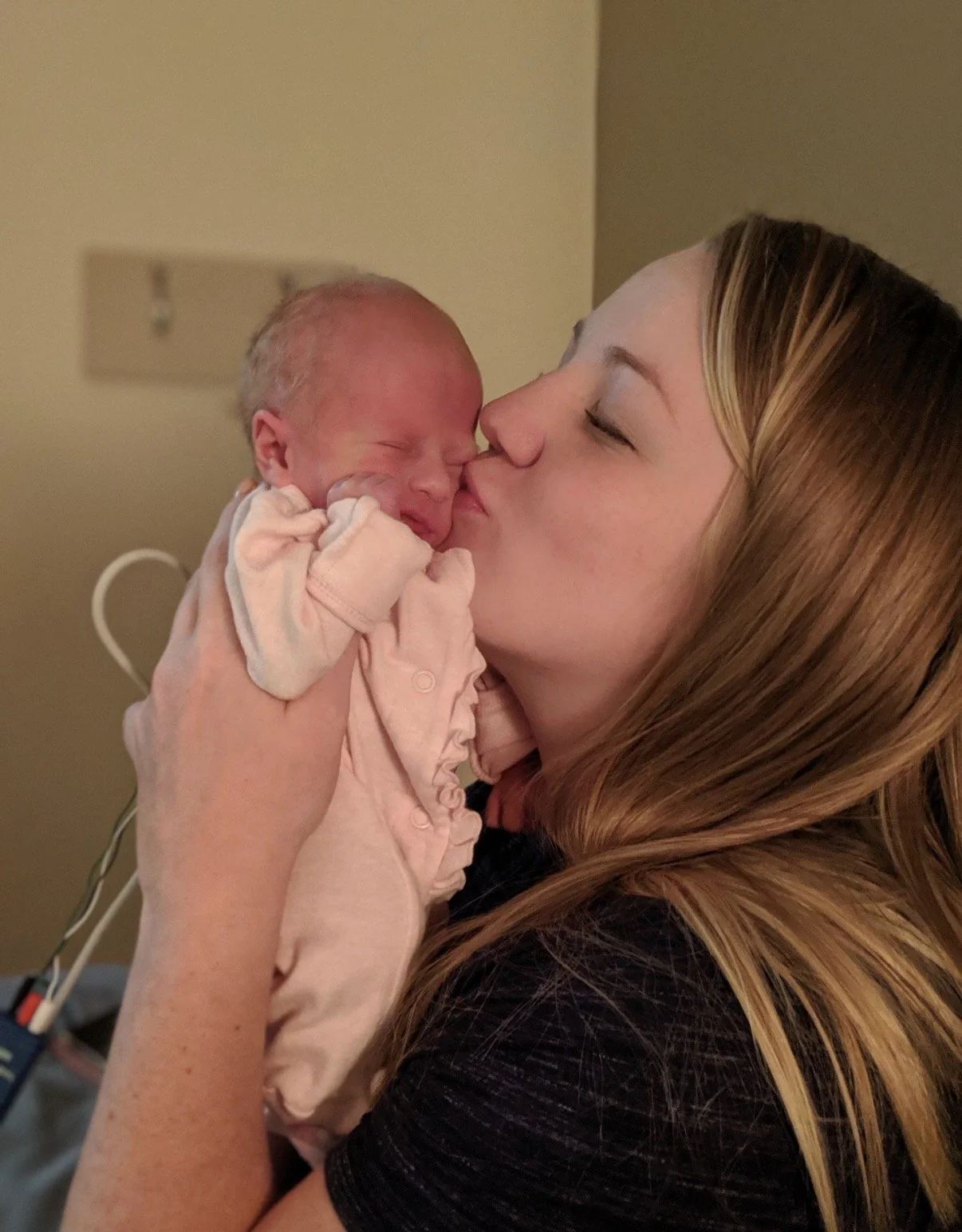 A woman holding a newborn baby and giving a gentle kiss on the baby's nose, in a hospital room.