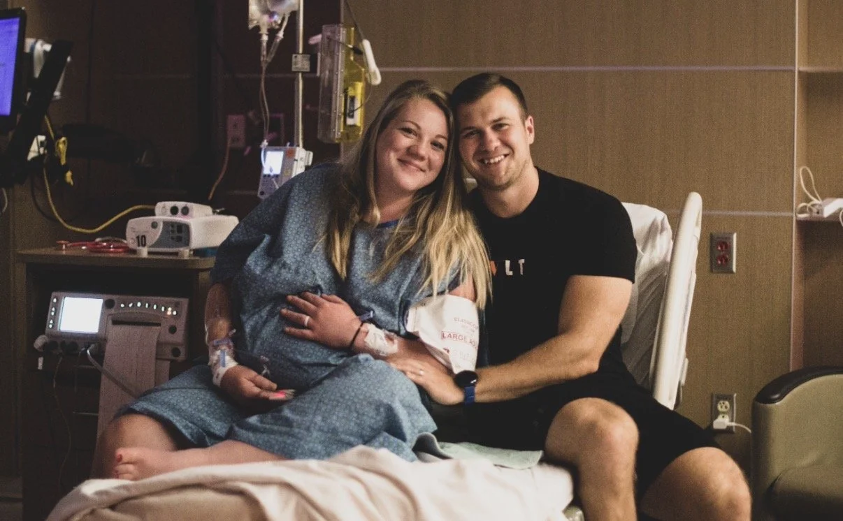A woman in a hospital gown sitting on a hospital bed holding her hand with an IV line, smiling and leaning toward a man who is sitting on the bed next to her, also smiling. The background shows medical equipment and monitors.