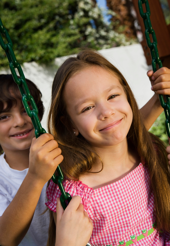 Two displaced children in Georgia relative care , a girl with long brown hair and a boy with dark hair, on a swing set outdoors. The girl is smiling at the camera, and the boy is partially behind her, also smiling.