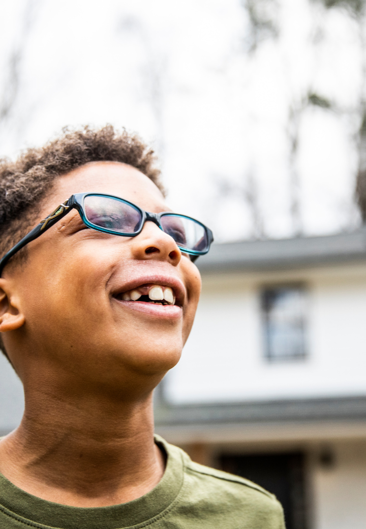 Smiling boy in foster care with curly hair and sunglasses outside, with house and trees in the background.