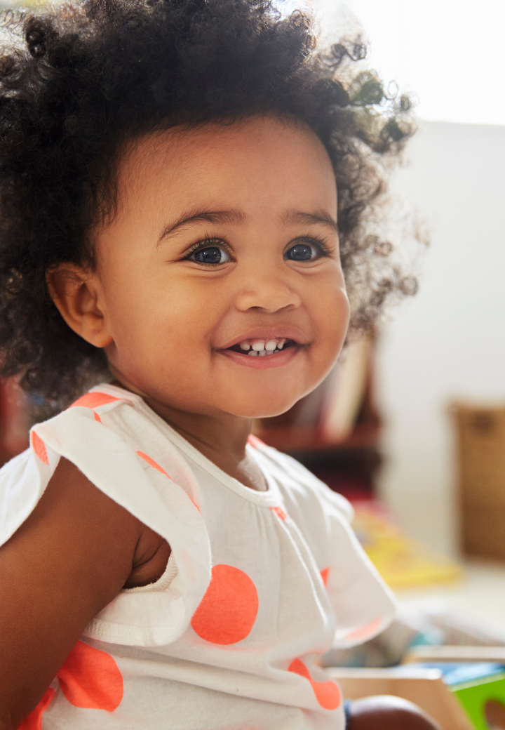 Close-up of a smiling young displaced toddler in relative care with curly hair and big blue eyes, wearing a white shirt with orange polka dots.