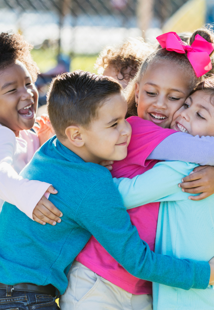 Displaced Children in kinship care wearing colorful clothing hugging and smiling outdoors on a sunny day.