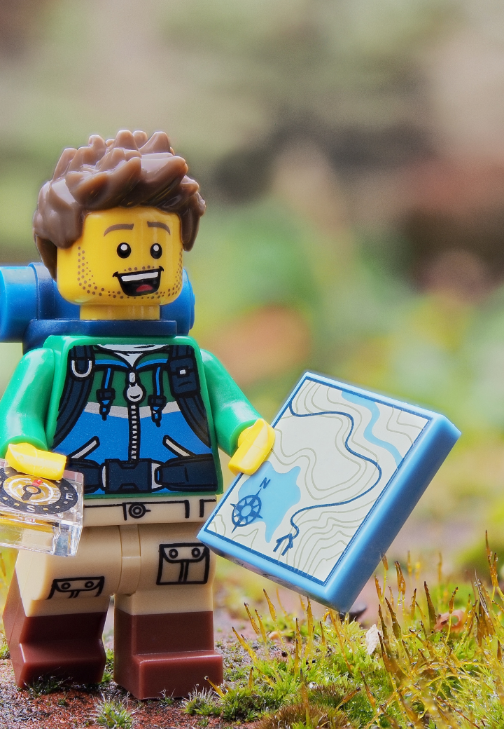A LEGO figurine of a male hiker with a green jacket, tan pants, and a blue backpack, holding a map and a compass, standing outdoors on green moss and grass with blurred trees in the background.