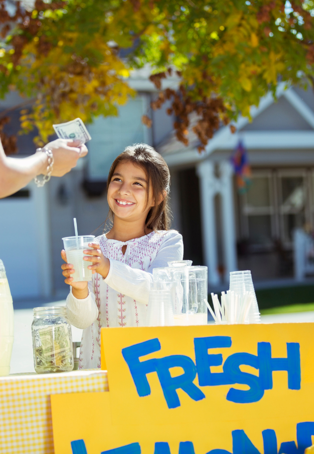 A young girl in your community at a lemonade stand holding a cup of lemonade, smiling, with a person handing her a dollar bill, under a tree with green and yellow leaves, in a suburban neighborhood.