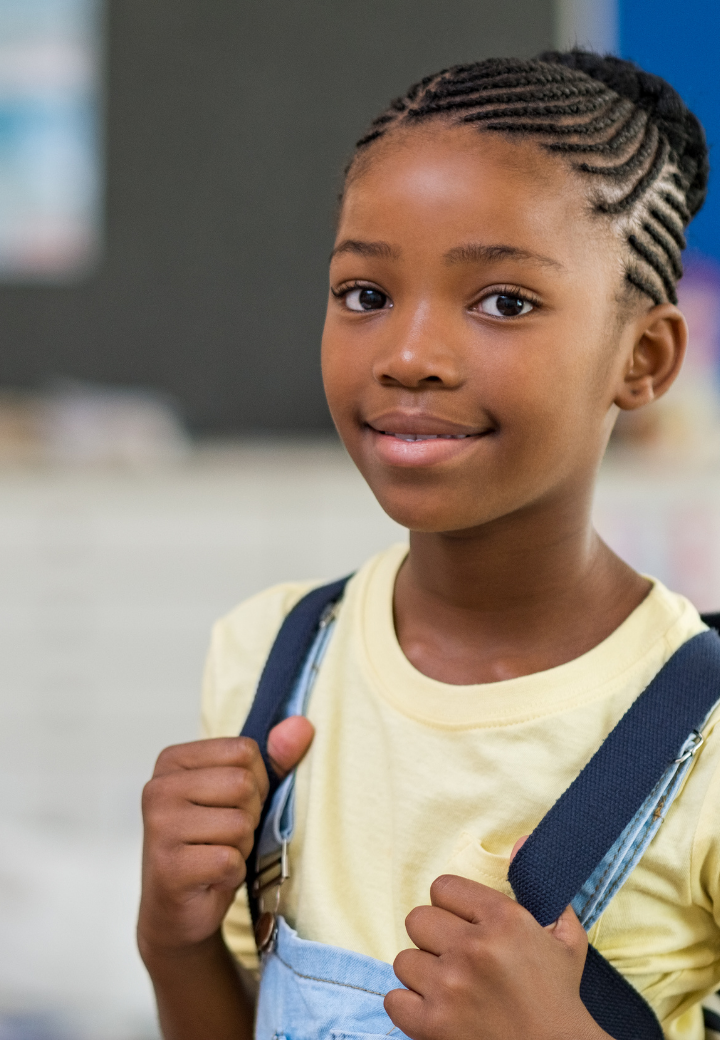 A young girl displaced in Georgia grands raising grands relative care with braided hair wearing a yellow shirt and carrying a backpack, smiling confidently at the camera.