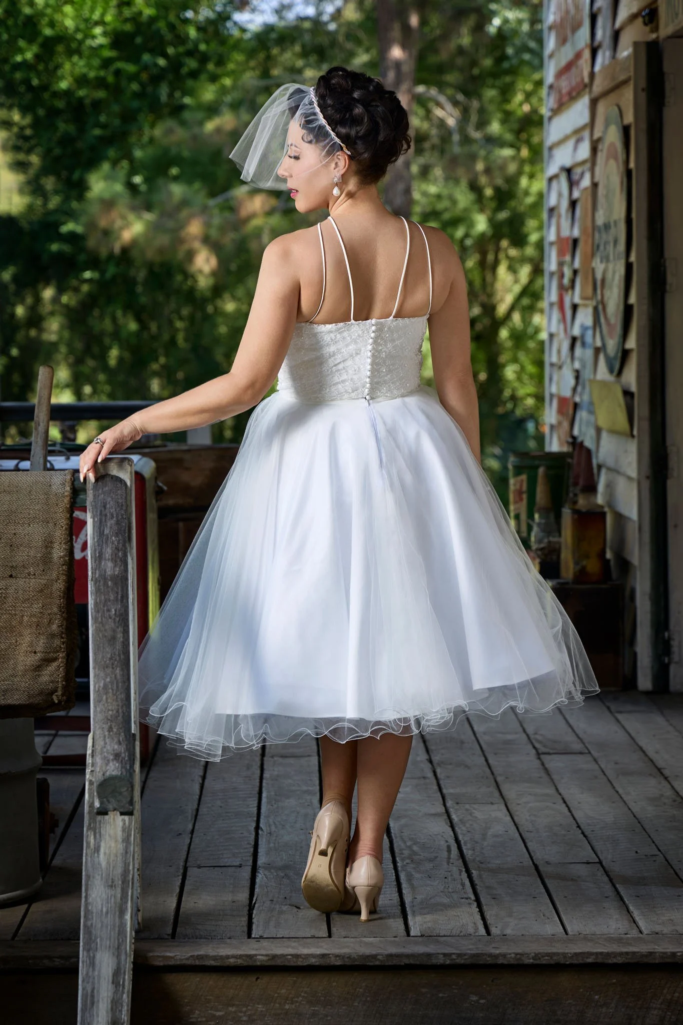 A woman in a white wedding dress standing on a wooden porch, touching a railing, with trees in the background and a rustic building on her right. Image by JK Bridal vintage style wedding dresses.