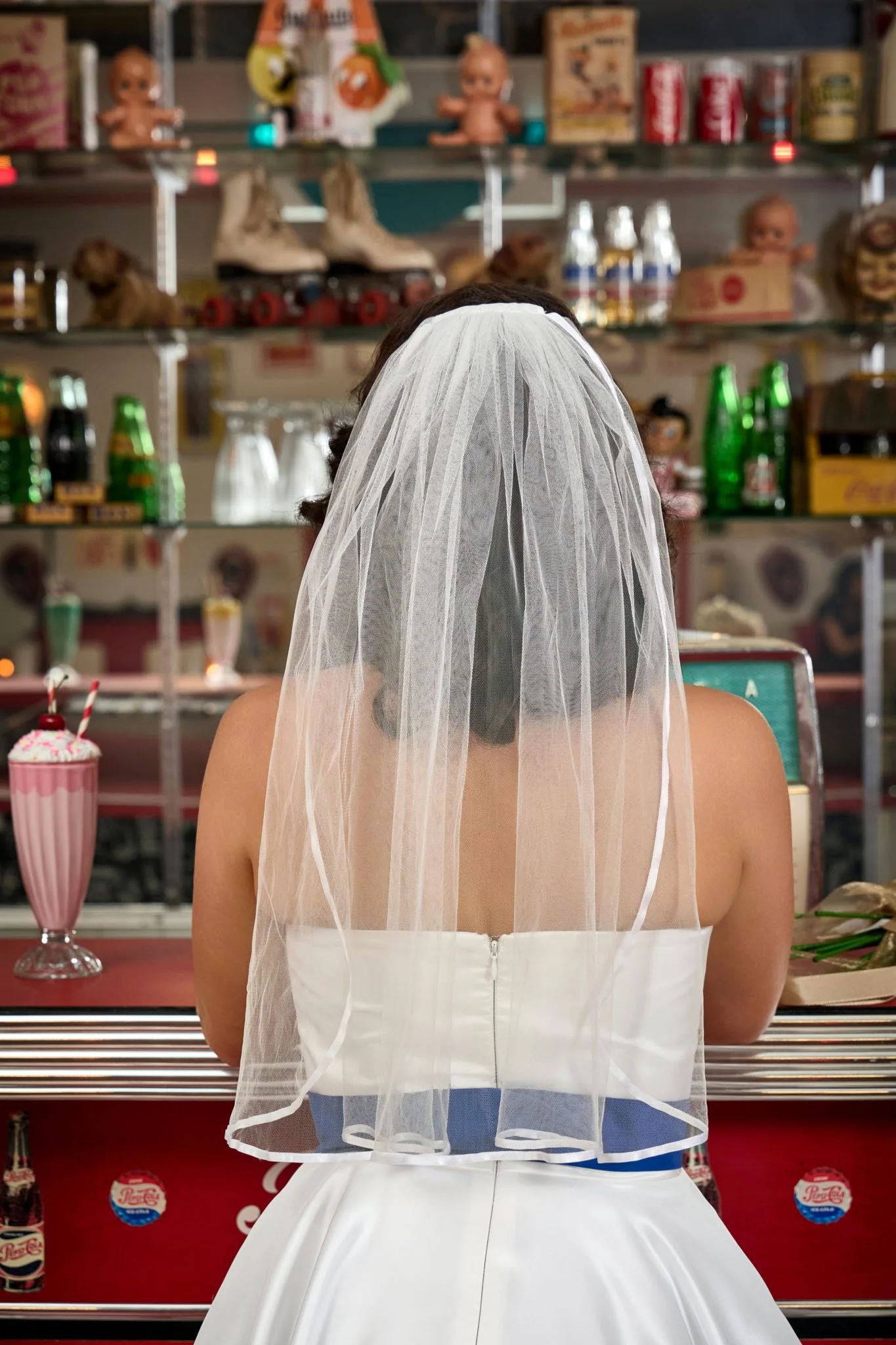 Person dressed in a white strapless dress with a veil, sitting at a counter in a vintage ice cream parlor with various vintage toys, bottles, and snacks on shelves in the background. Image by JK Bridal vintage style wedding dresses.