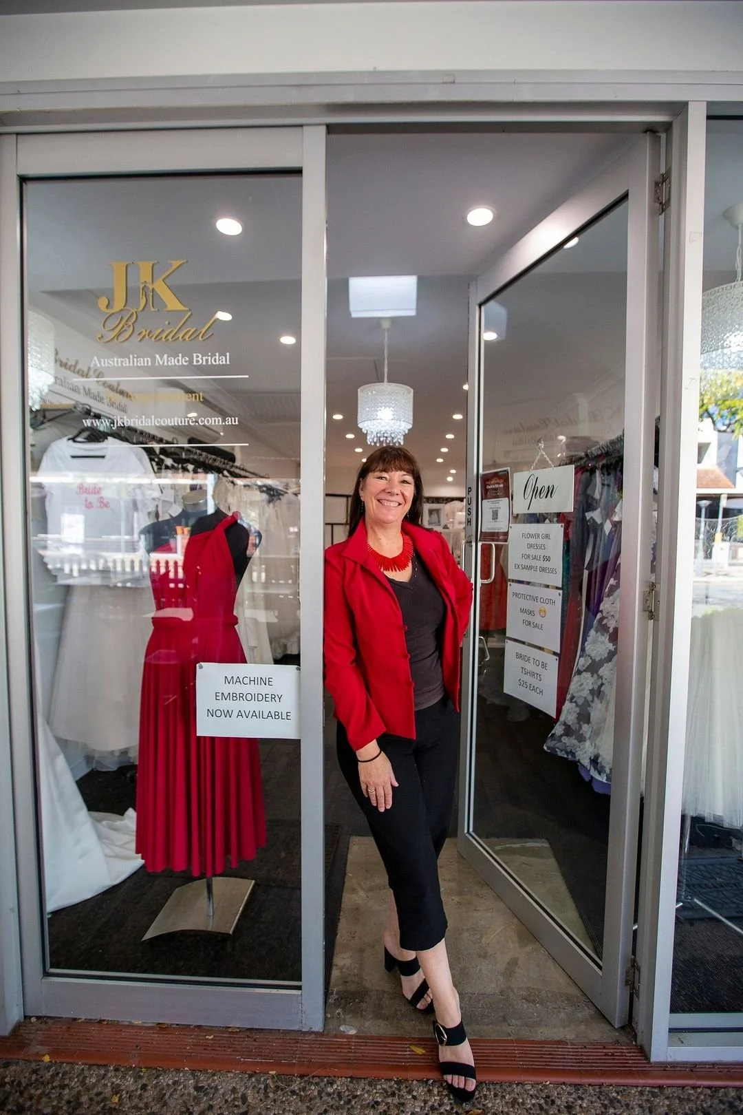 Woman dressed in black pants and a red jacket standing in front of a bridal shop with dresses on display and signs advertising sale items. Image by JK Bridal vintage style wedding dresses.