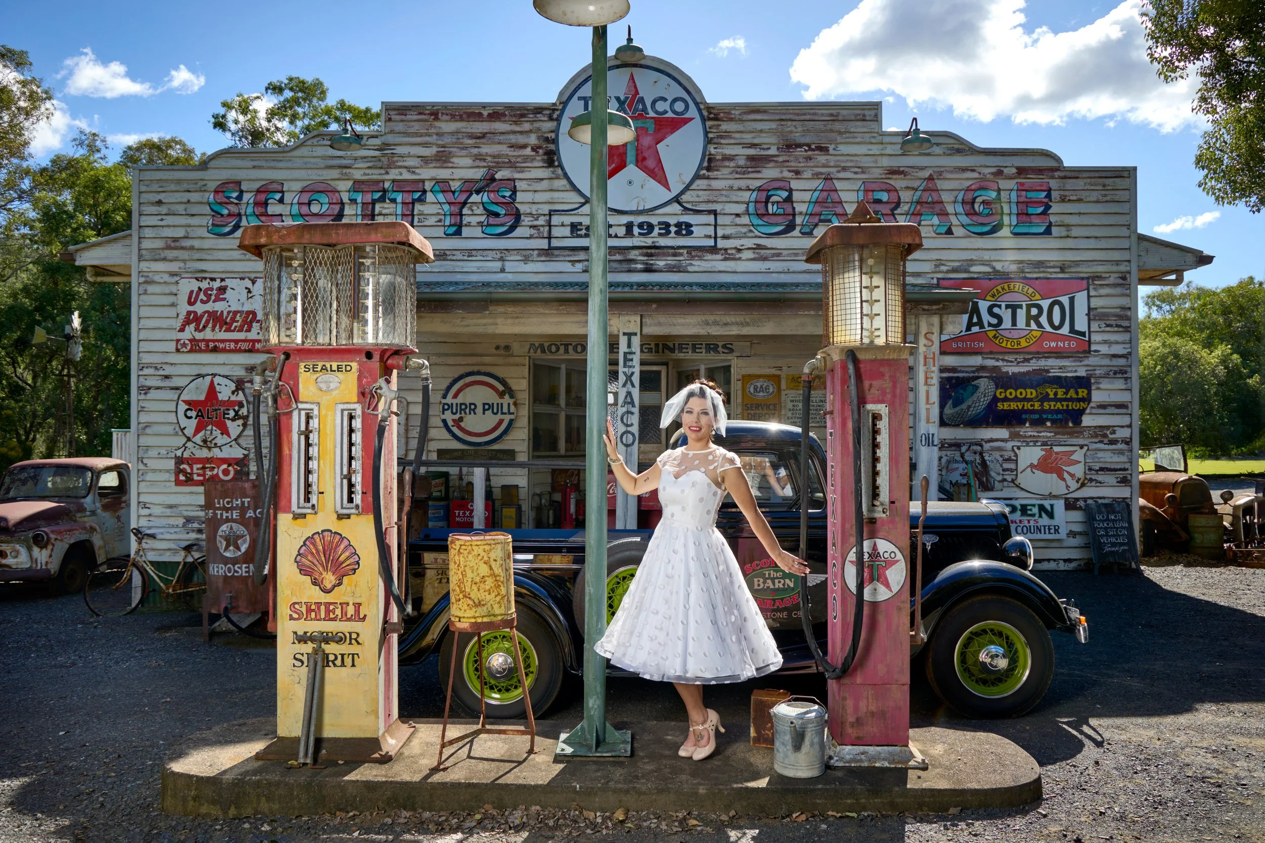 A woman in a vintage white dress standing between two old gas pumps at a retro gas station, with vintage cars and signs in the background. Image by JK Bridal vintage style wedding dresses.