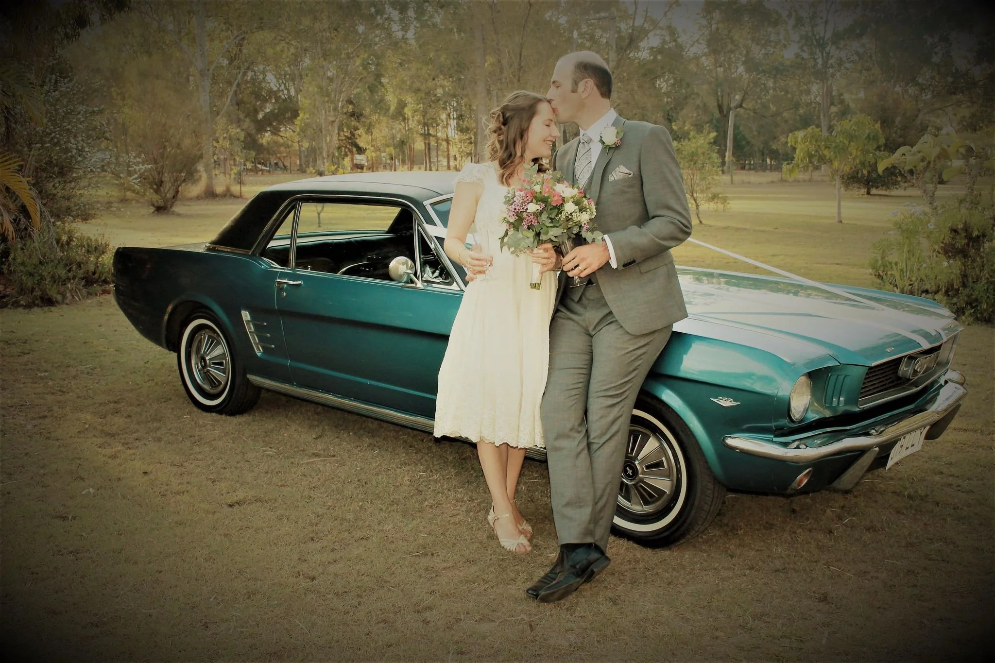 A bride and groom in wedding attire lean against a vintage green Ford Mustang, smiling and sharing a kiss in an outdoor park with trees in the background. Image by JK Bridal vintage style wedding dresses.
