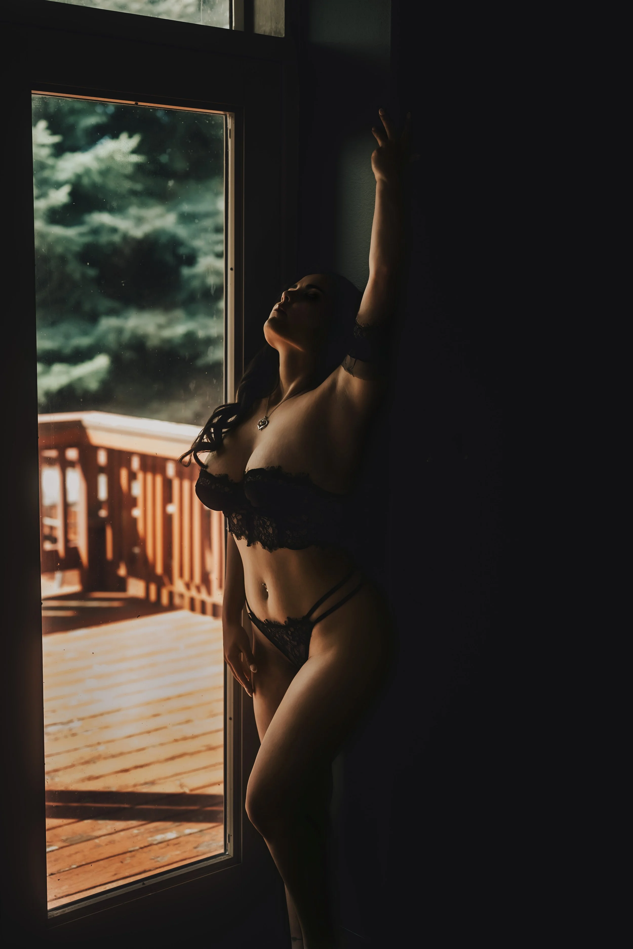 A woman in black lace lingerie stands by a sliding glass door, with her eyes closed and her left arm raised, touching the wall. Sunlight streams through the door, casting shadows inside. A wooden deck and green trees are visible outside.
