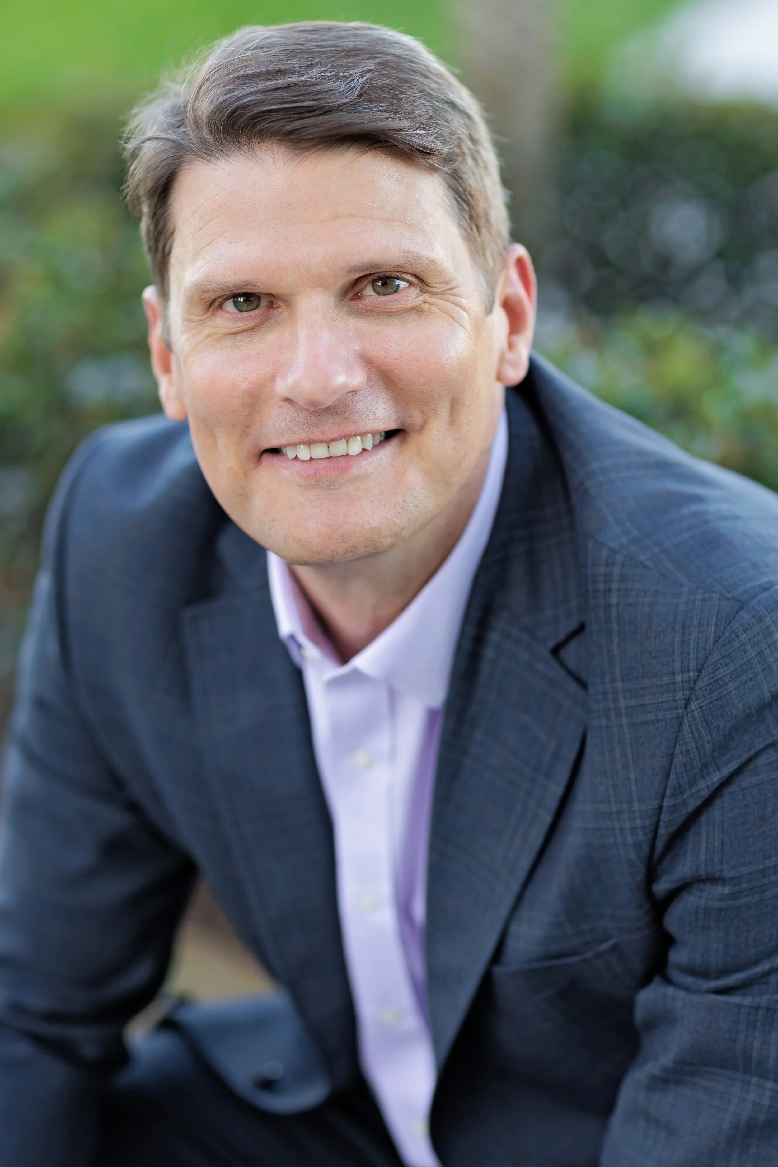 Close-up of a man in a dark blue checkered suit and white shirt, smiling outdoors with greenery in the background.