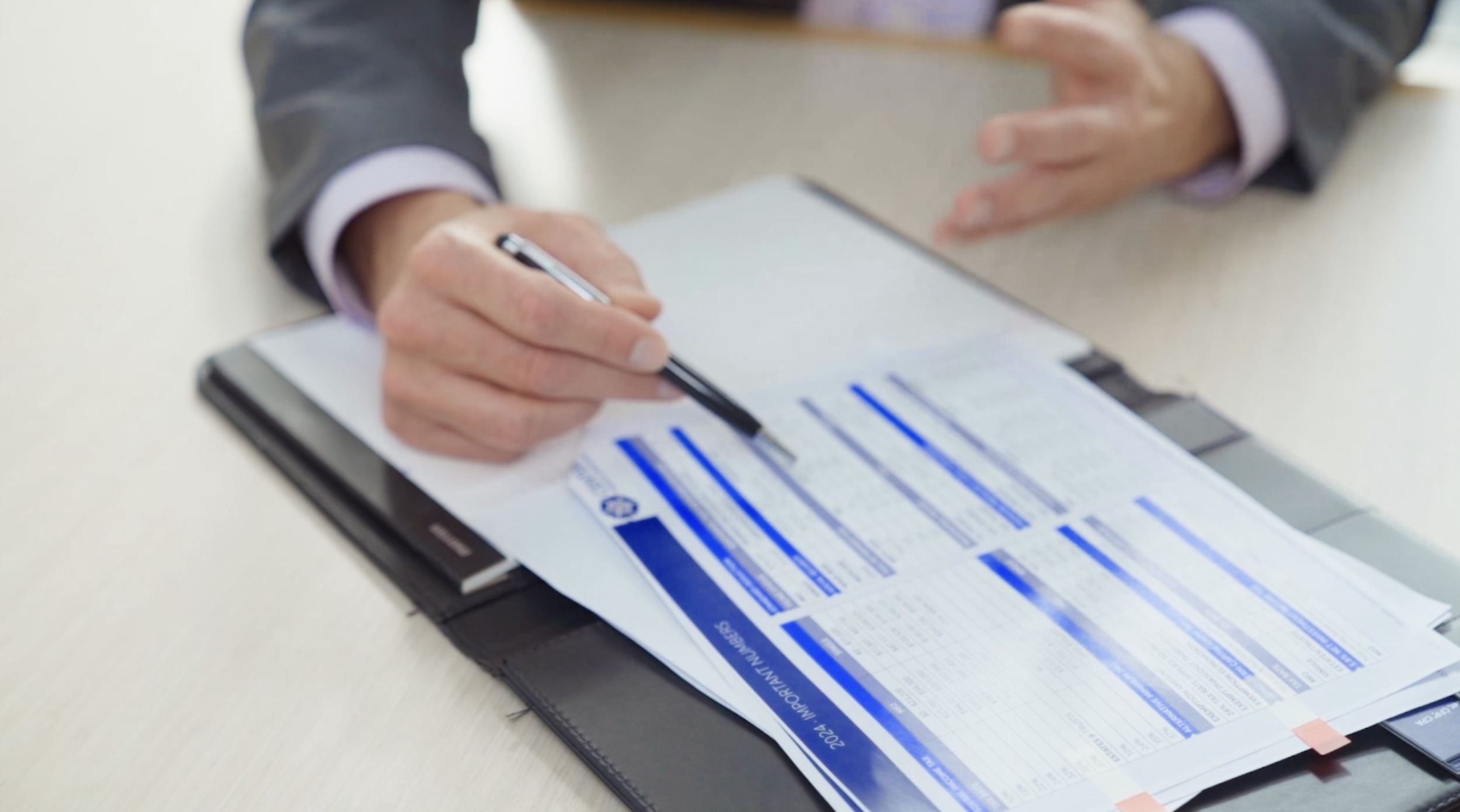 Person in a suit holding a pen, filling out a form on a clipboard on a white table.
