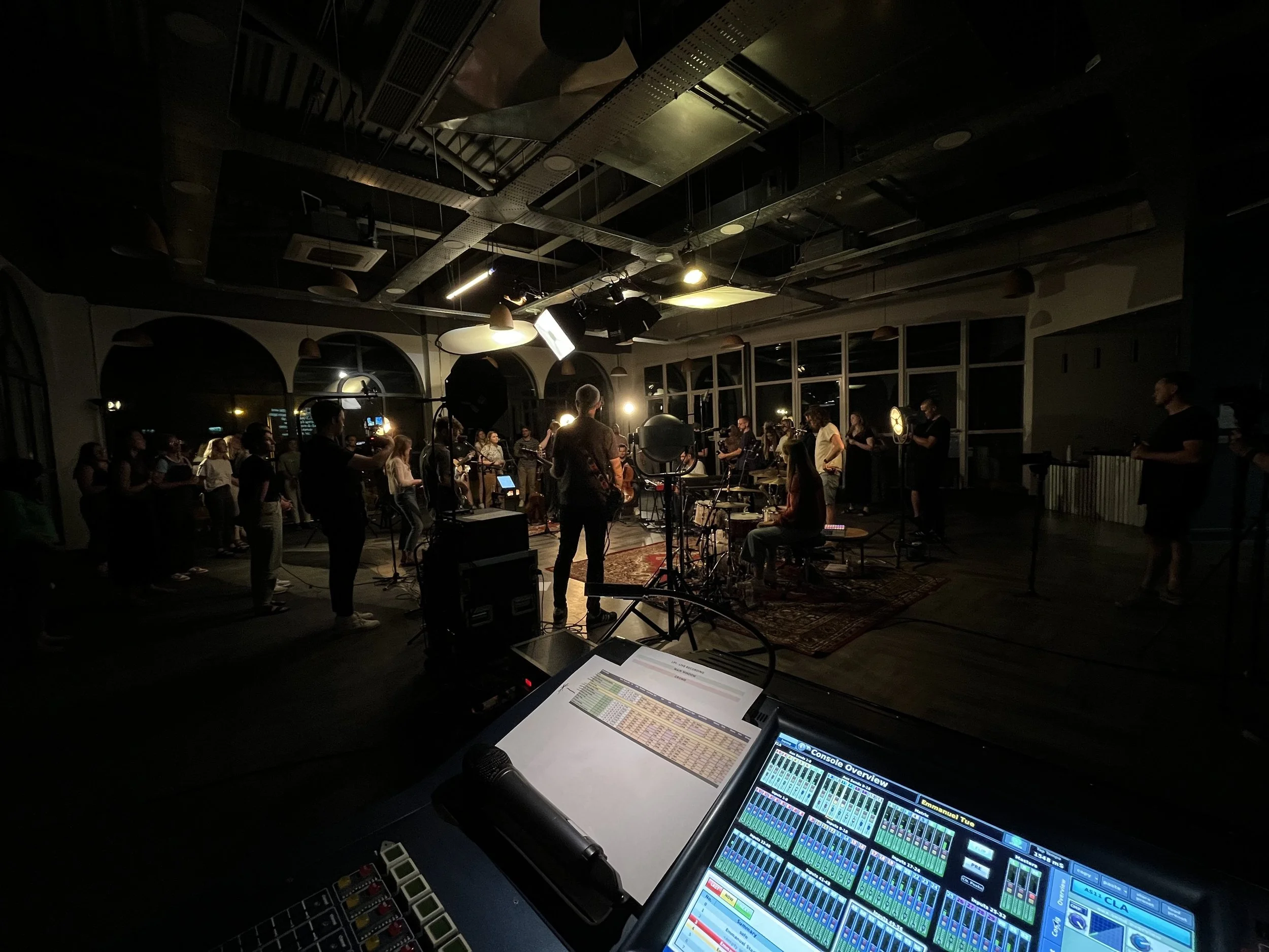 A music concert or performance taking place indoors with musicians and performers gathered on stage, illuminated by stage lighting. Audience members are seated and standing, watching the show, with some recording or taking photos. The view is from the perspective of the sound or lighting technician, looking at a mixing console and control equipment in the foreground.