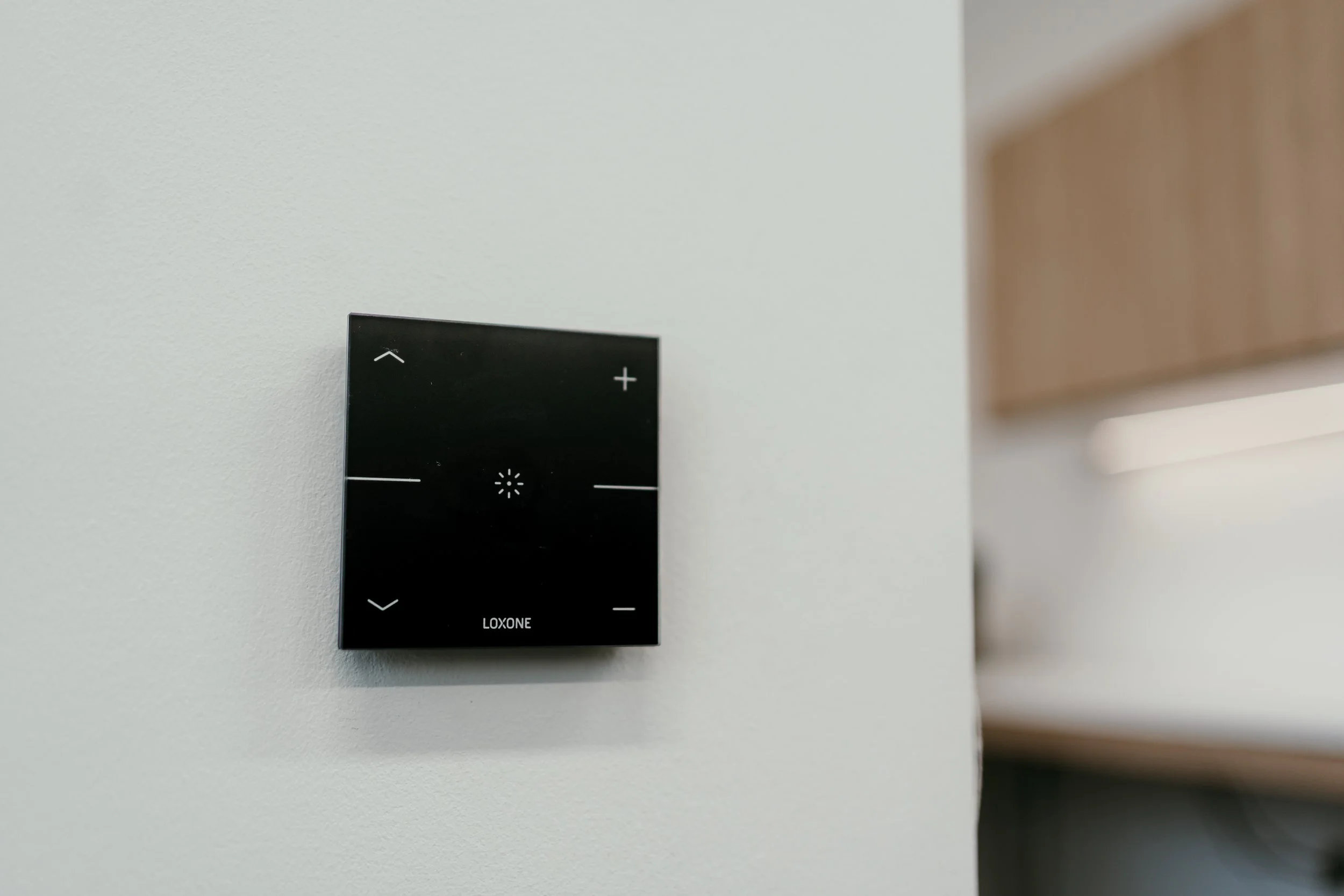 A black Loxone glass-plated switch with up and down arrows on the left, a lighting symbol in the middle, and plus and minus symbols on the right