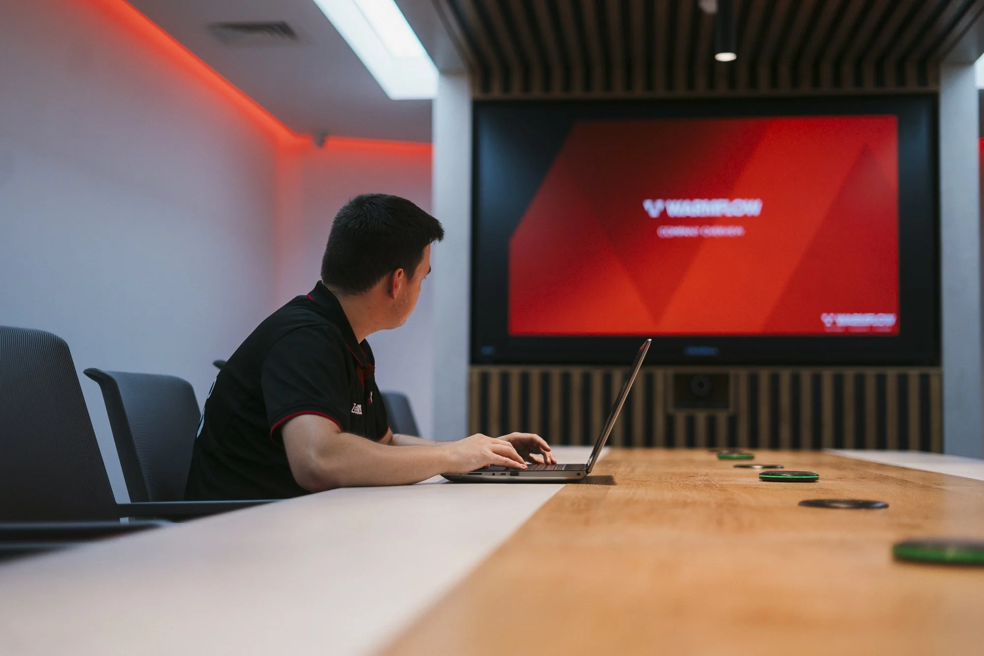 A man in a black polo shirt typing on a laptop looks away from us at a display screen in a conference room