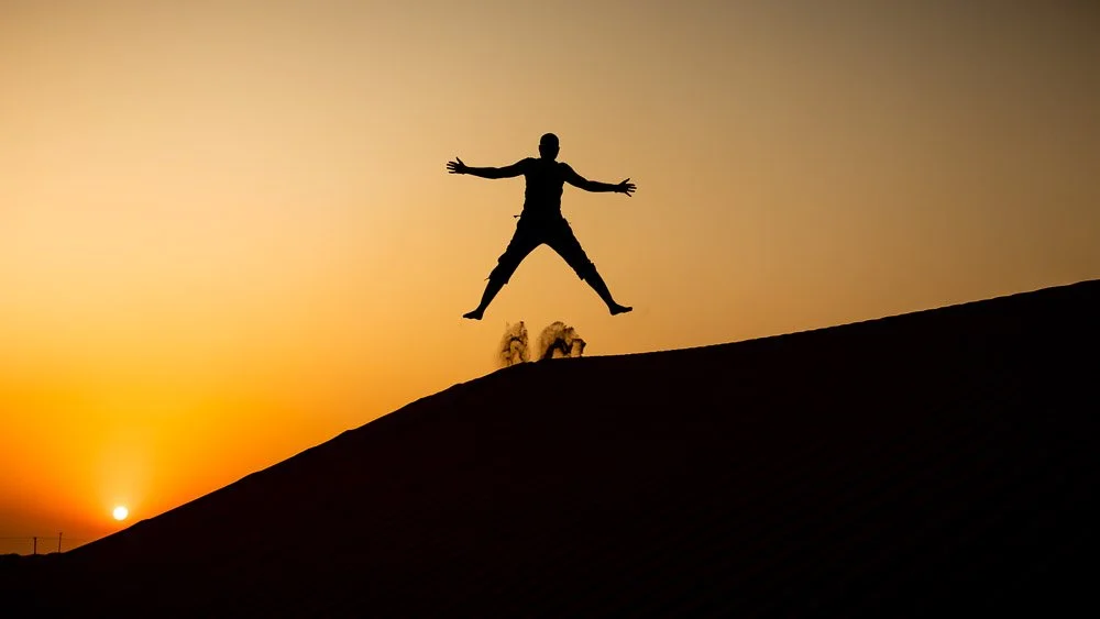 A figure suspended in a leap above a dune at sunset