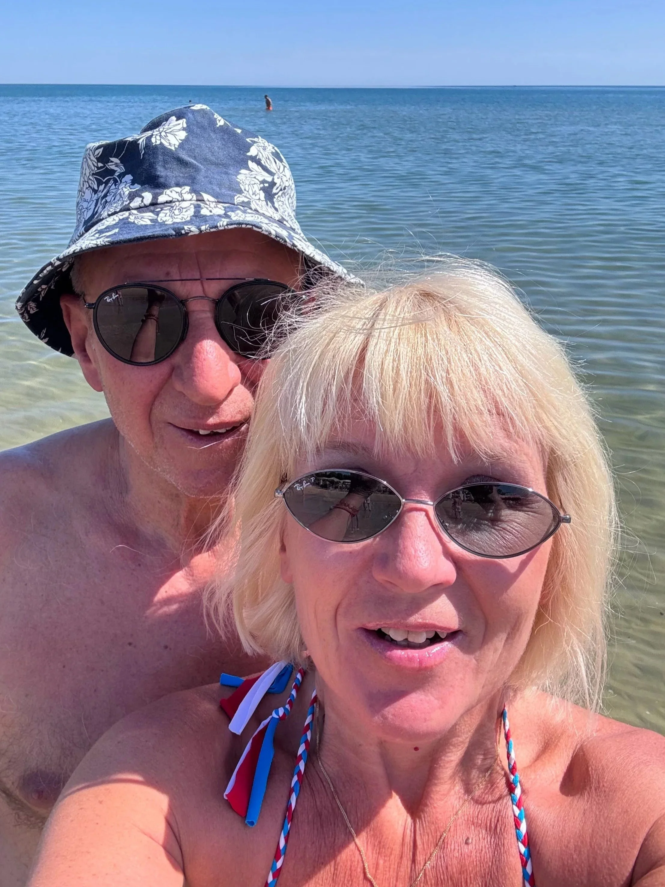 Anita and Umberto in a selfie on a beach with the blue waves of the sea in the background.