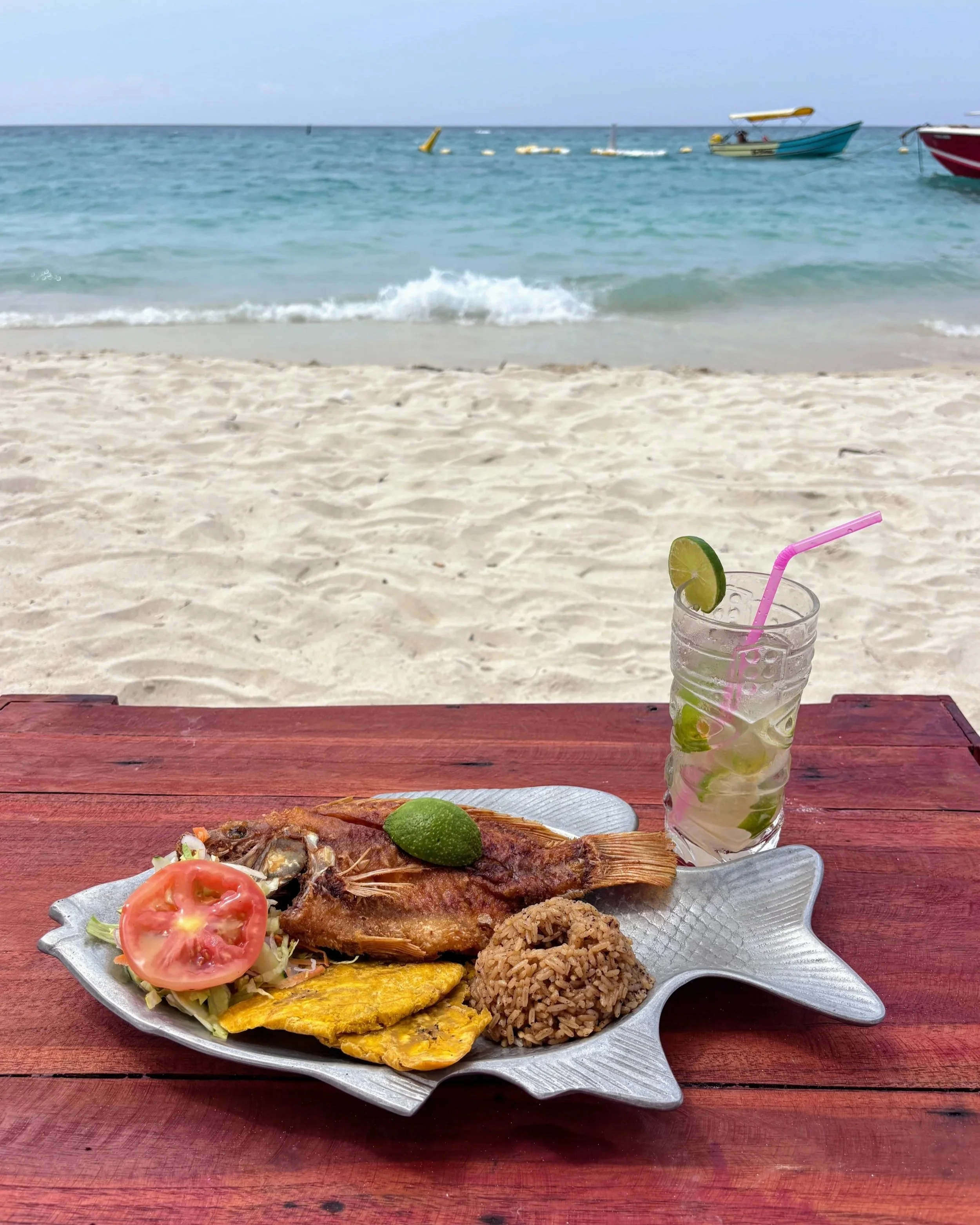 A lunch dish of grilled fish, a mojito cocktail, on a wooden table on a white sand beach with turquoise water and  boats in the background