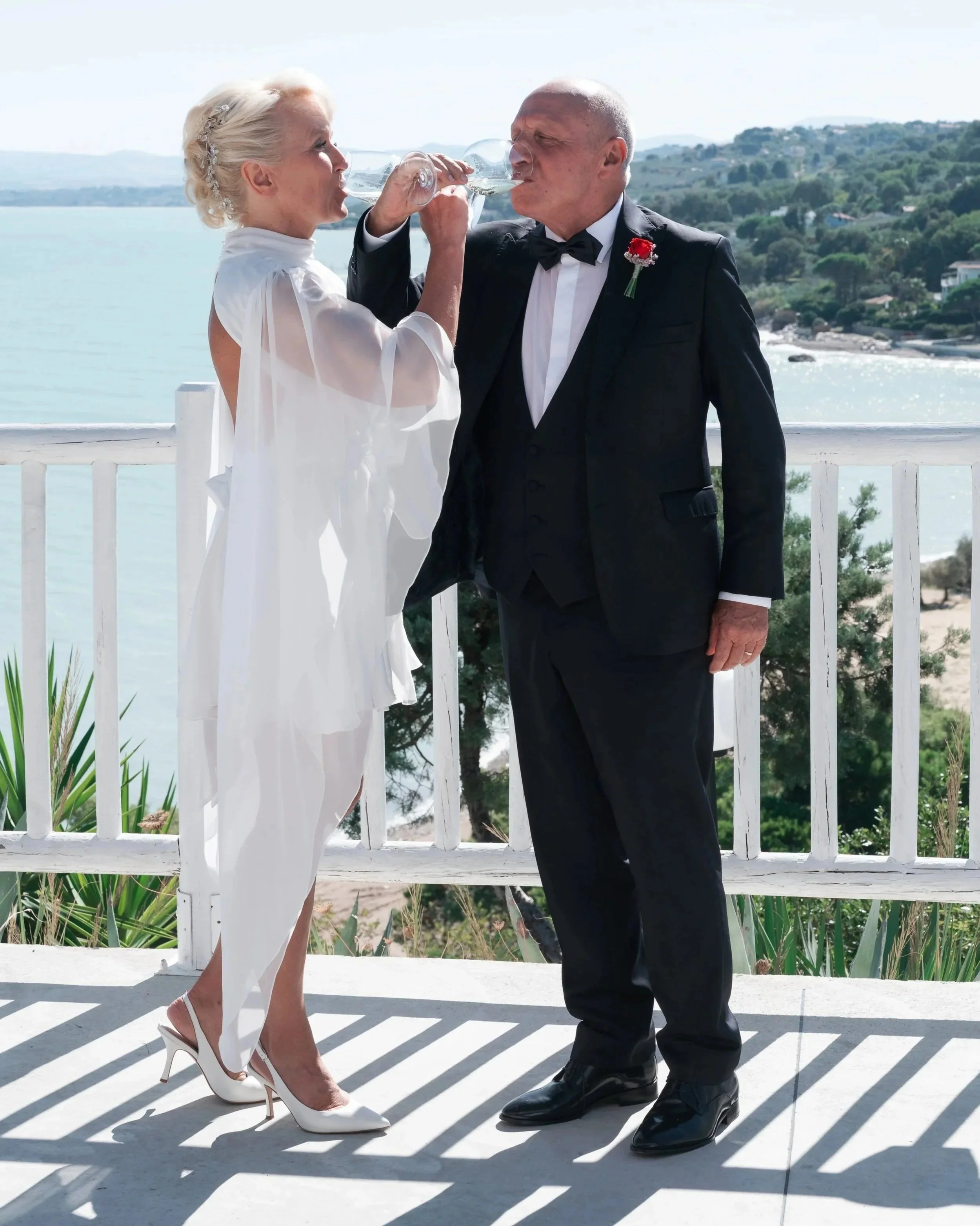 anita and umberto just married raising a toast with their arms crossed, with the Adriatic sea in the background