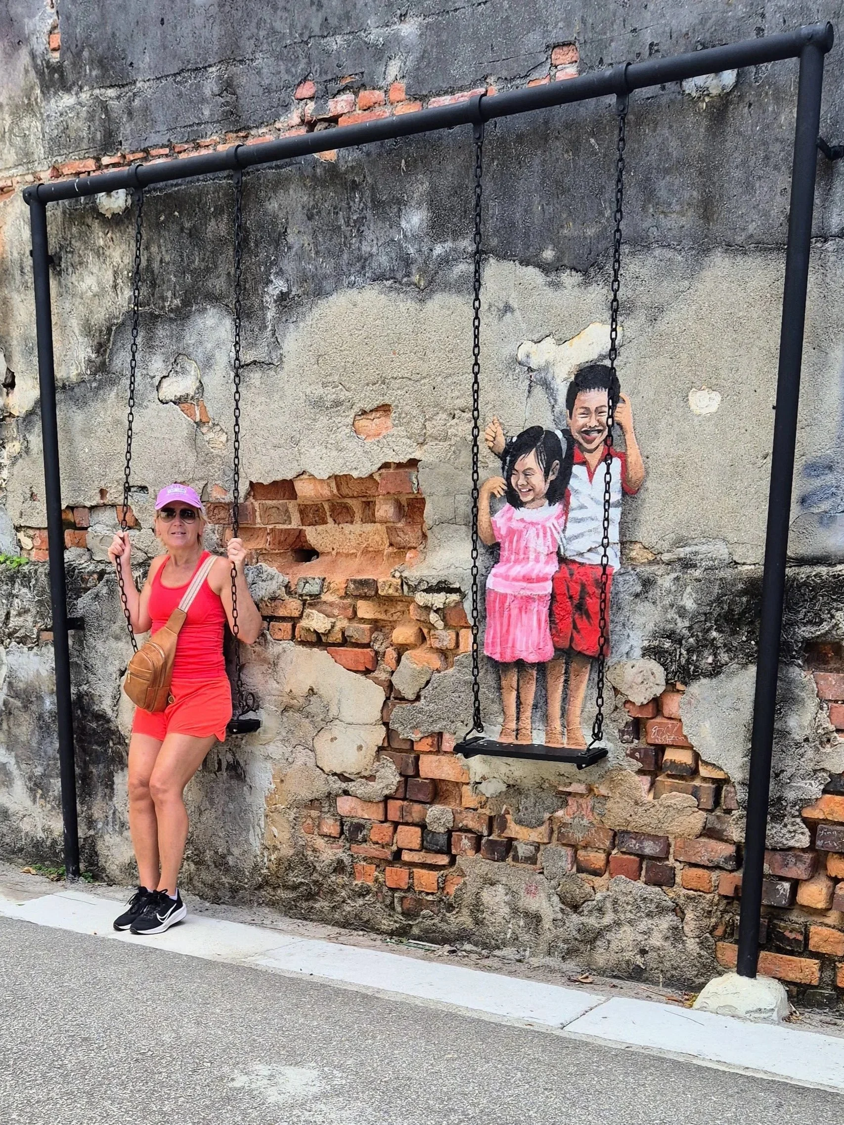 Anita in front of a mural featuring two swings and two Chinese children on the other swing. The photo was taken in George Town, Penang, Malaysia.