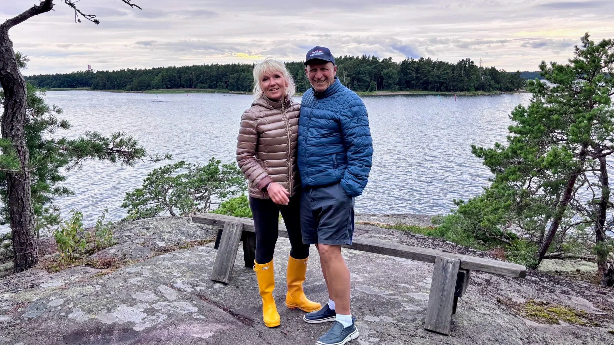 Anita and Umberto on a rocky island in the Finnish archipelago, with some trees around them and the sea and another forested island in the background.