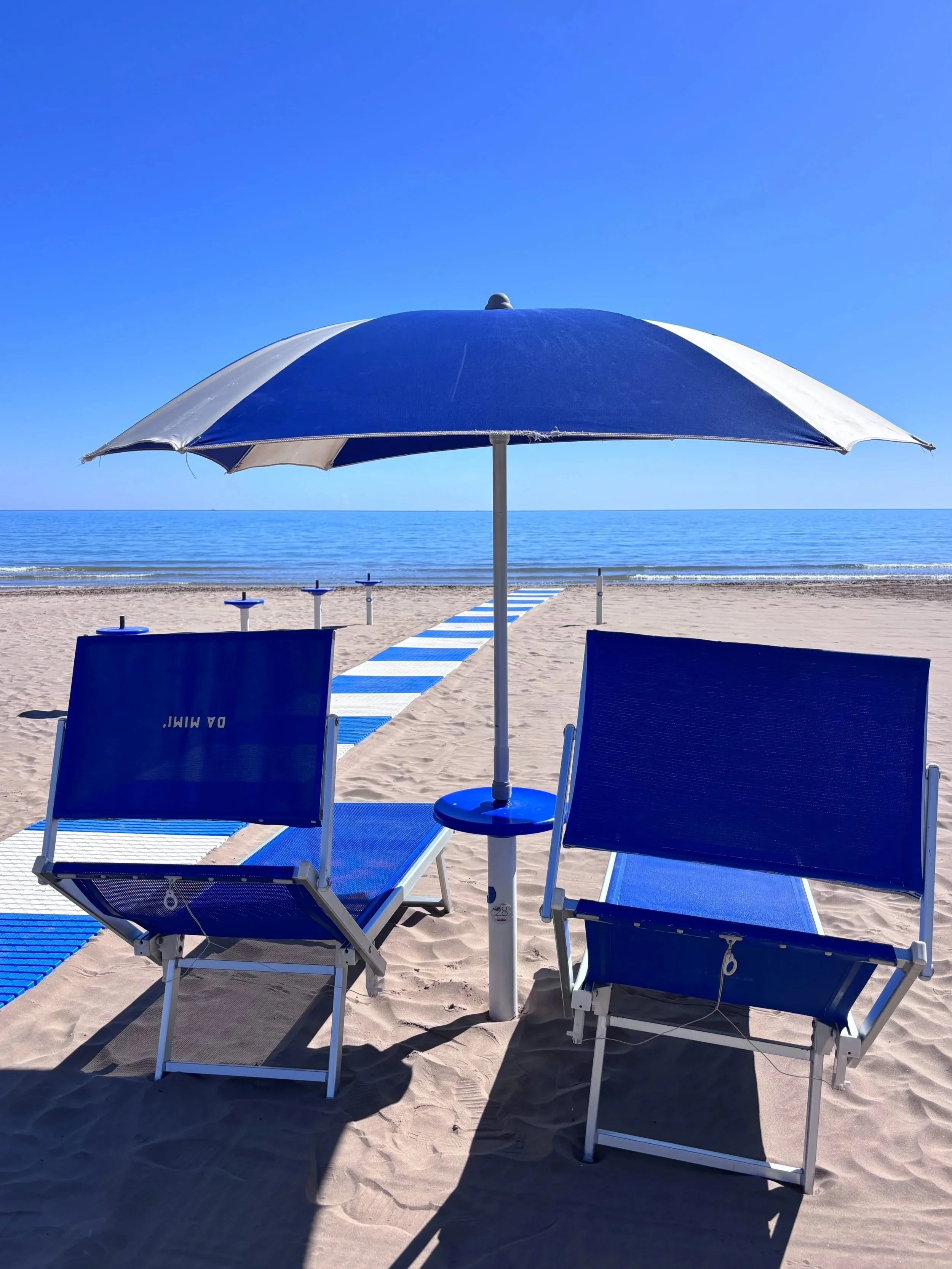 A beach umbrella with two sunbeds on a sandy beach with the blue sea in the background
